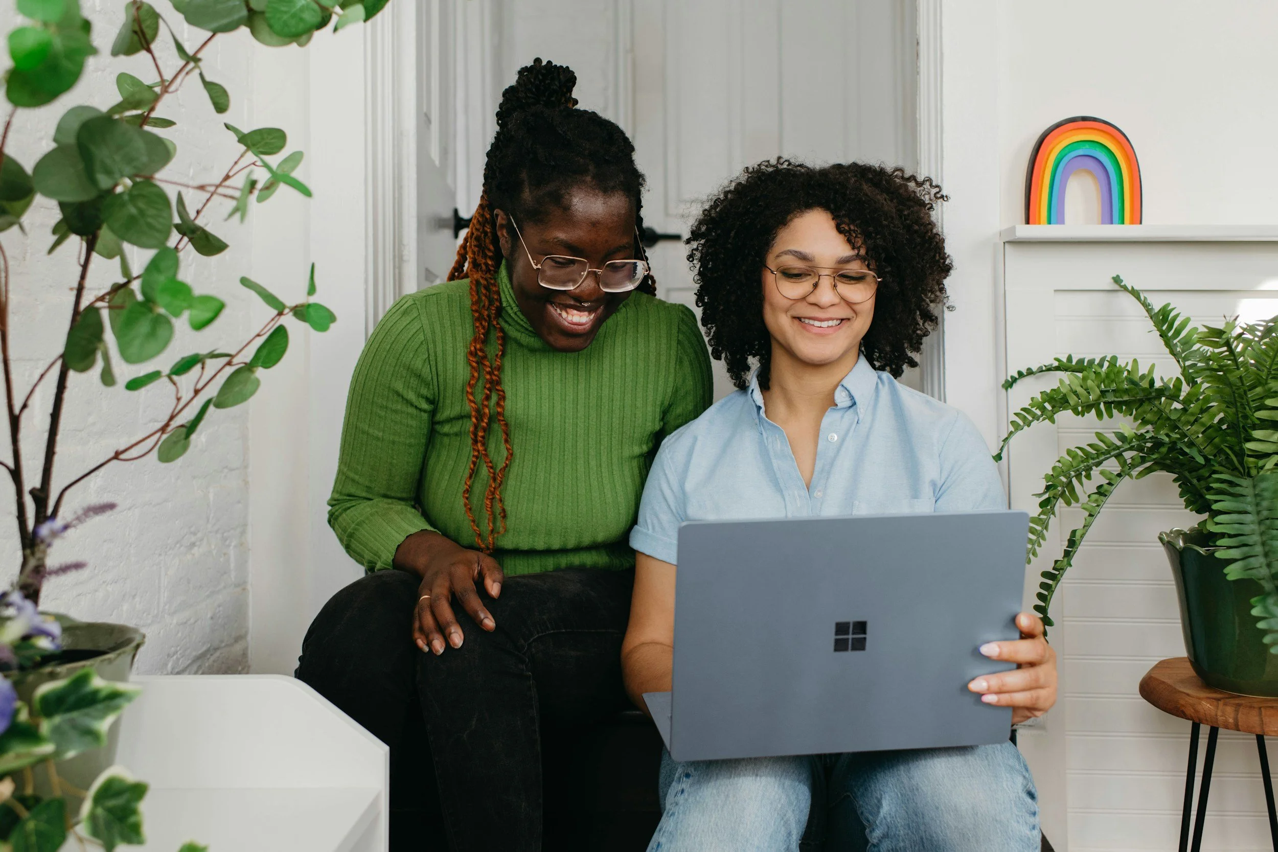 Two women sitting together, one showing the laptop screen to the other, surrounded by houseplants, with a rainbow decoration on the white wall behind them.
