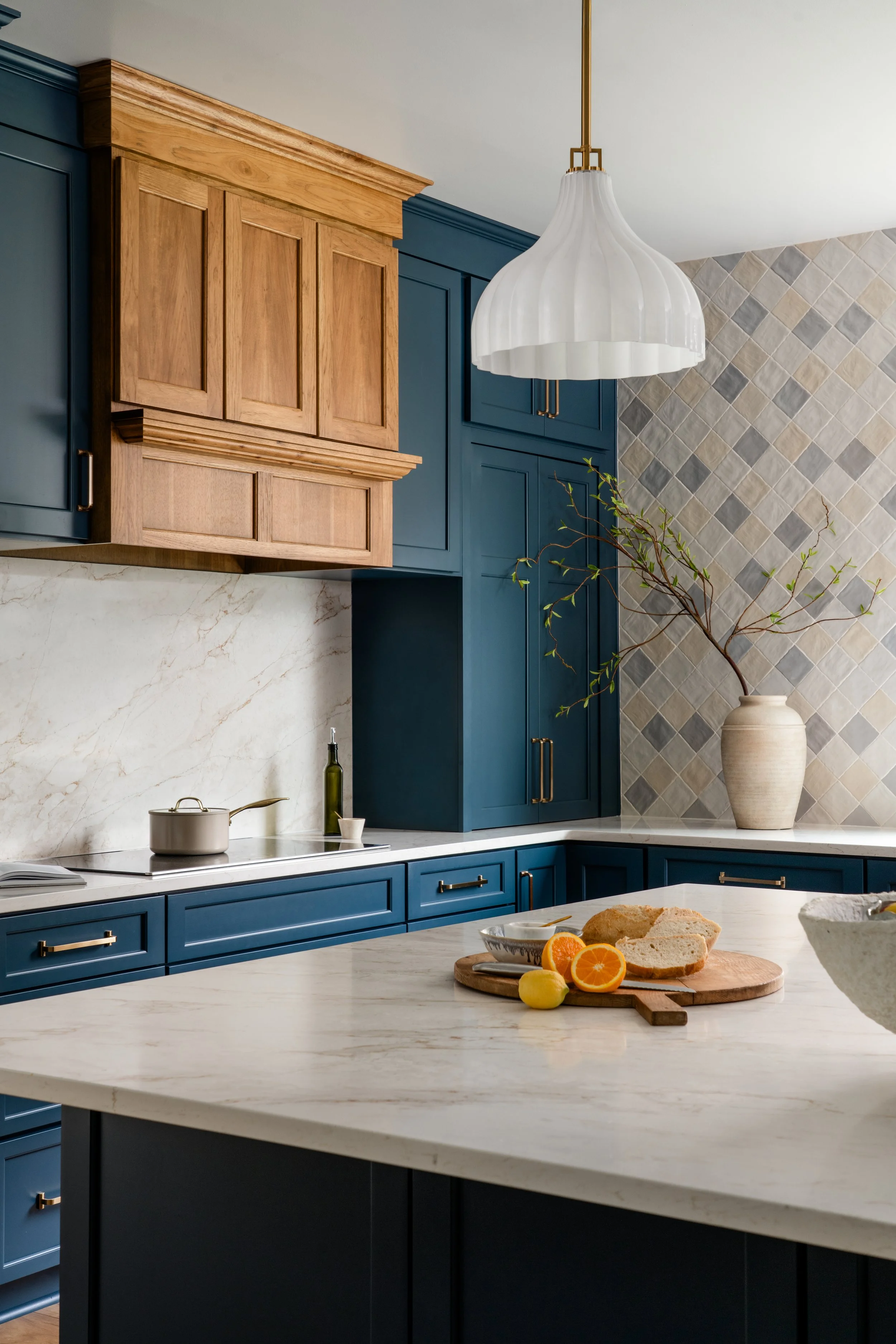 Modern kitchen with blue and wood cabinets, marble countertop, white pendant light, beige and gray tiled wall, large vase with branch, and some fruit and bread on the counter.