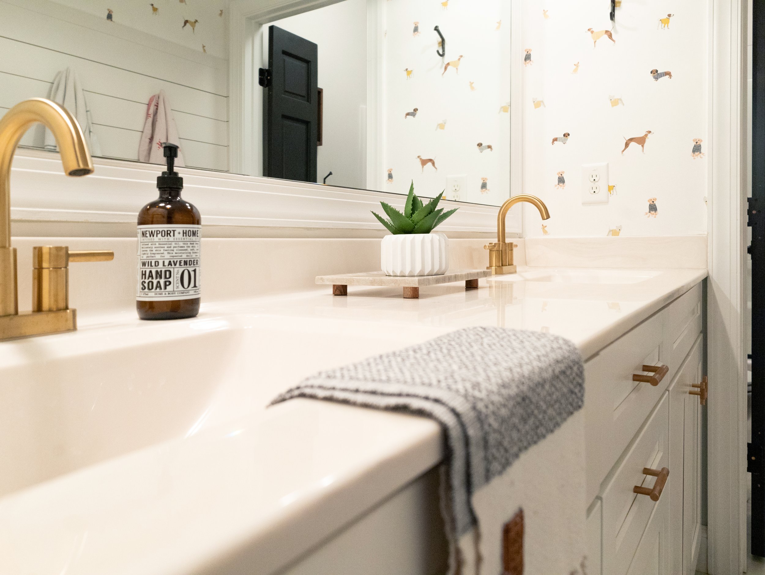 Bathroom vanity with a beige countertop, a gold faucet, a hand soap dispenser labeled 'Newport + Home Wild Lavender,' a potted succulent on a wooden tray, and a gray and white striped towel hanging from the edge. In the reflection of the mirror, a wall with wallpaper featuring small dogs, a black door, and some hanging towels.