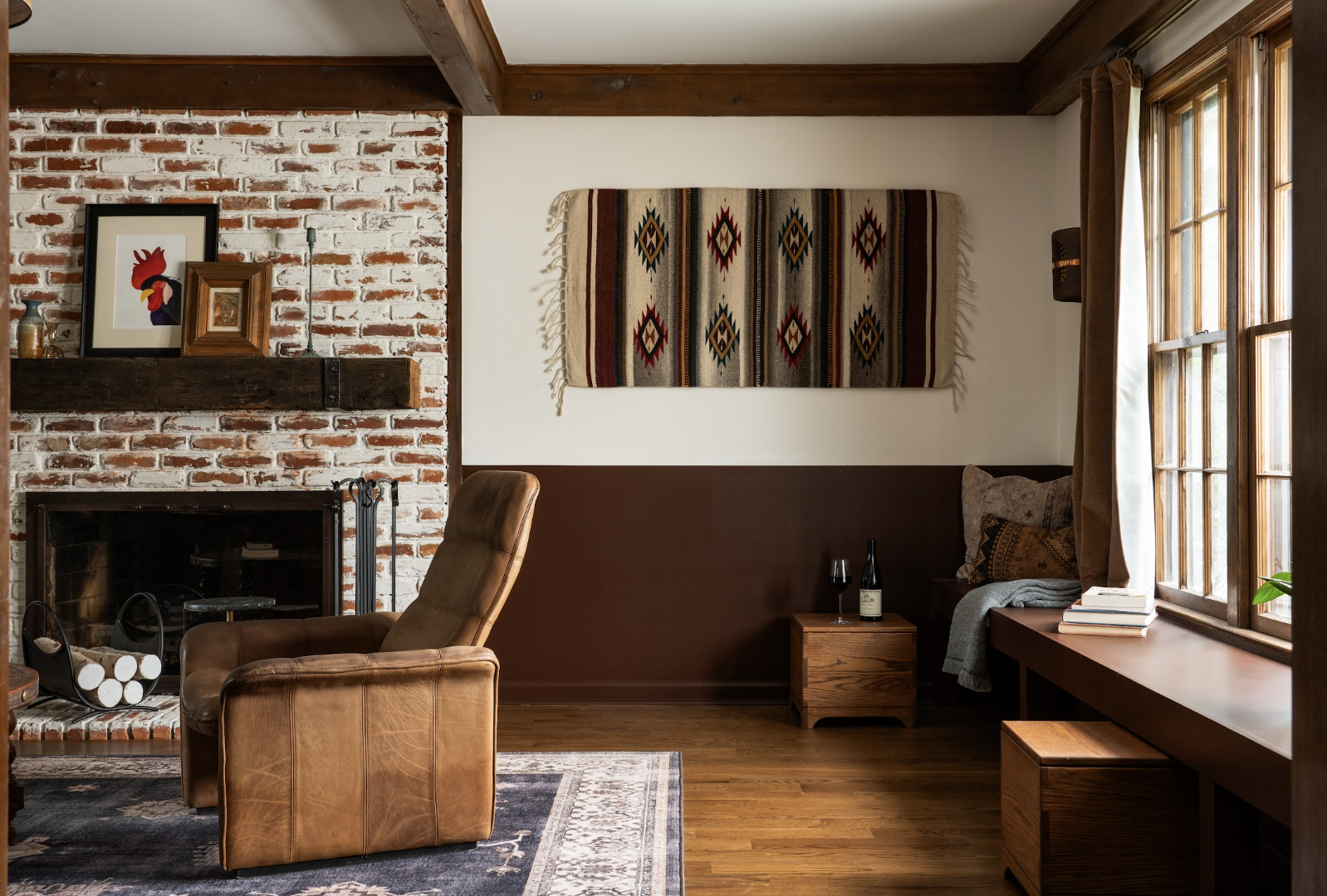 Living room with a brick fireplace, framed artwork, a leather armchair, a window seat with pillows and books, a woven wall hanging, and a window with brown curtains.