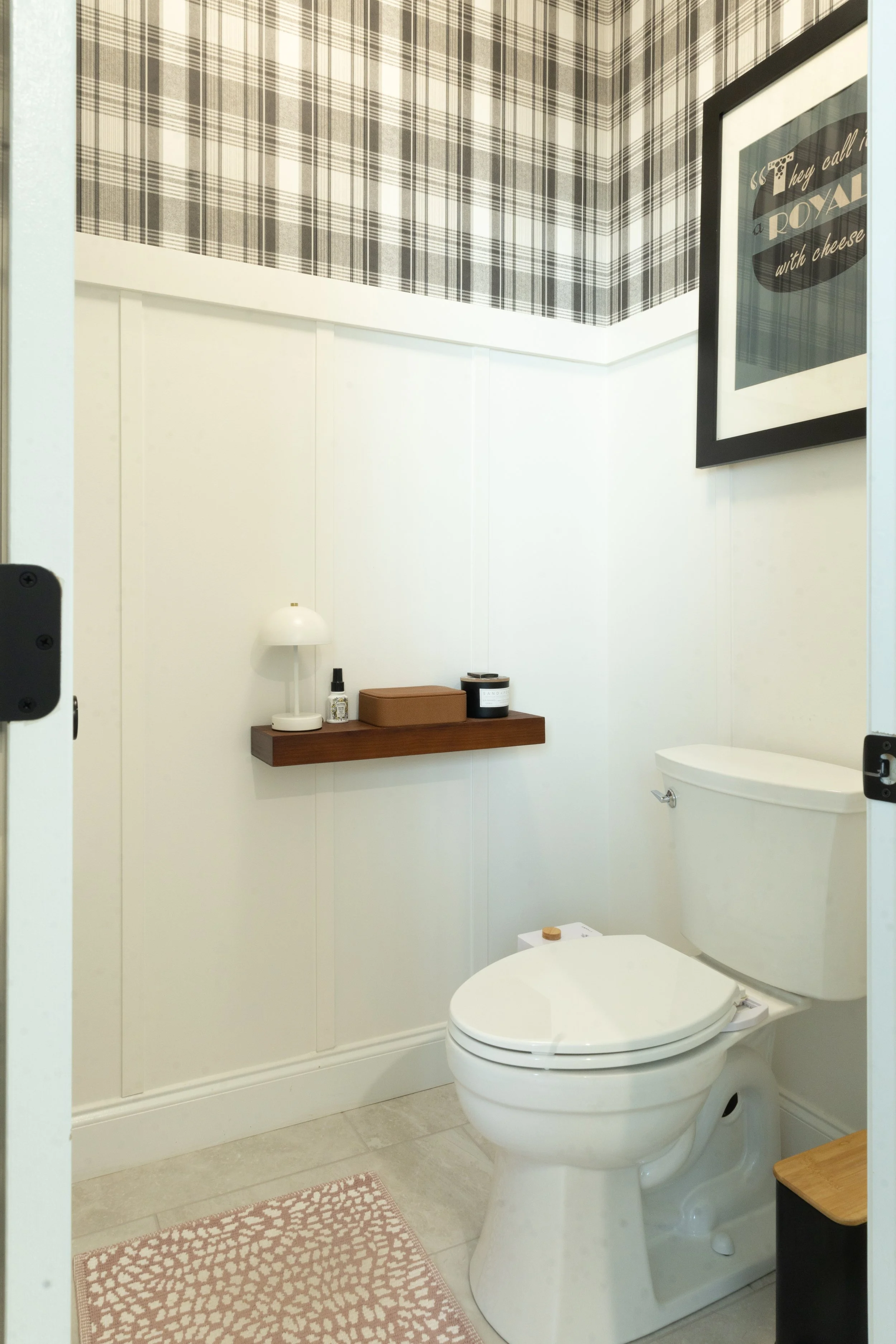 A small bathroom with white walls, a toilet, a wooden shelf with decorative items, and a framed picture on the wall.