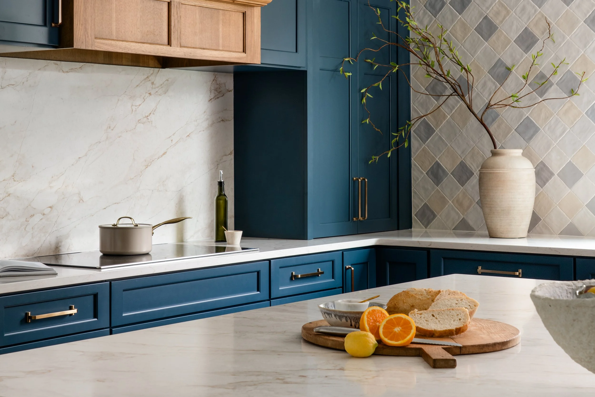 A modern kitchen with blue cabinetry, a white marble countertop, and a backsplash with grey tiles. A beige vase with branches and green leaves is on the counter. There is a pot, a bottle of oil, and a small cup on the counter. On the island, there is
