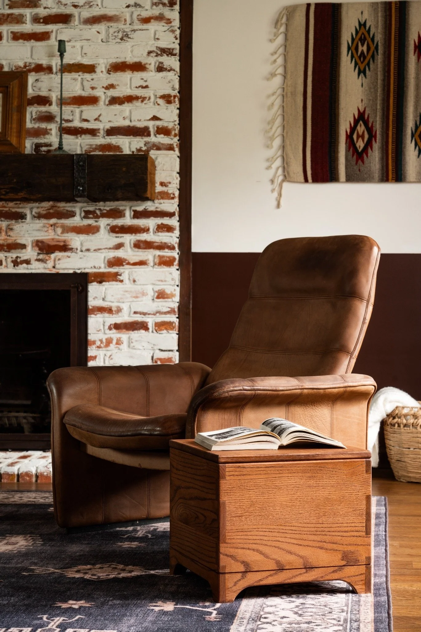 A cozy living room corner featuring a leather armchair, an open book on a wooden side table, a vintage brick fireplace with a wooden mantle, a patterned wall hanging, and a woven basket on the floor.