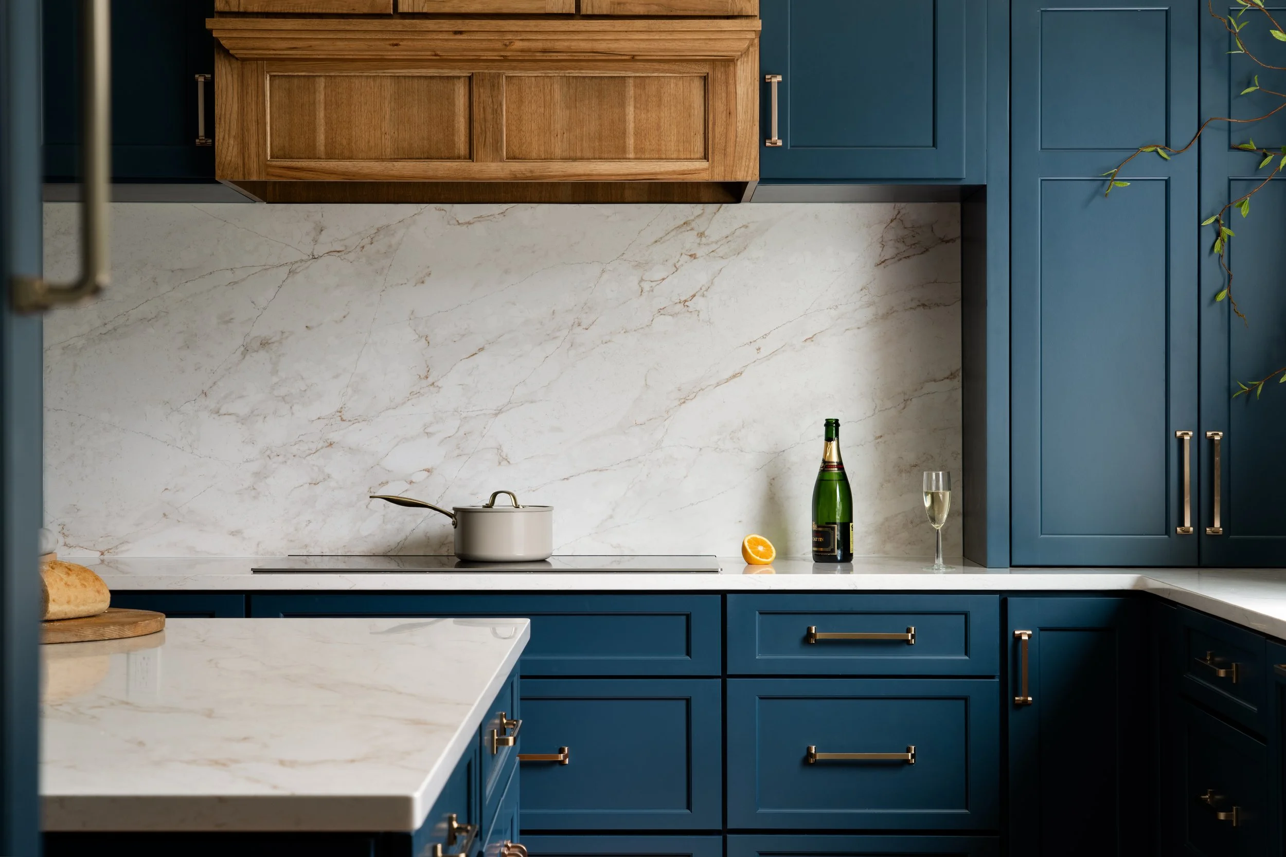 A modern kitchen with deep blue cabinets, a marble backsplash, and a white marble countertop. There is a white pot on the stovetop, a bottle of wine with a glass, a lemon half, and a piece of bread on a cutting board.