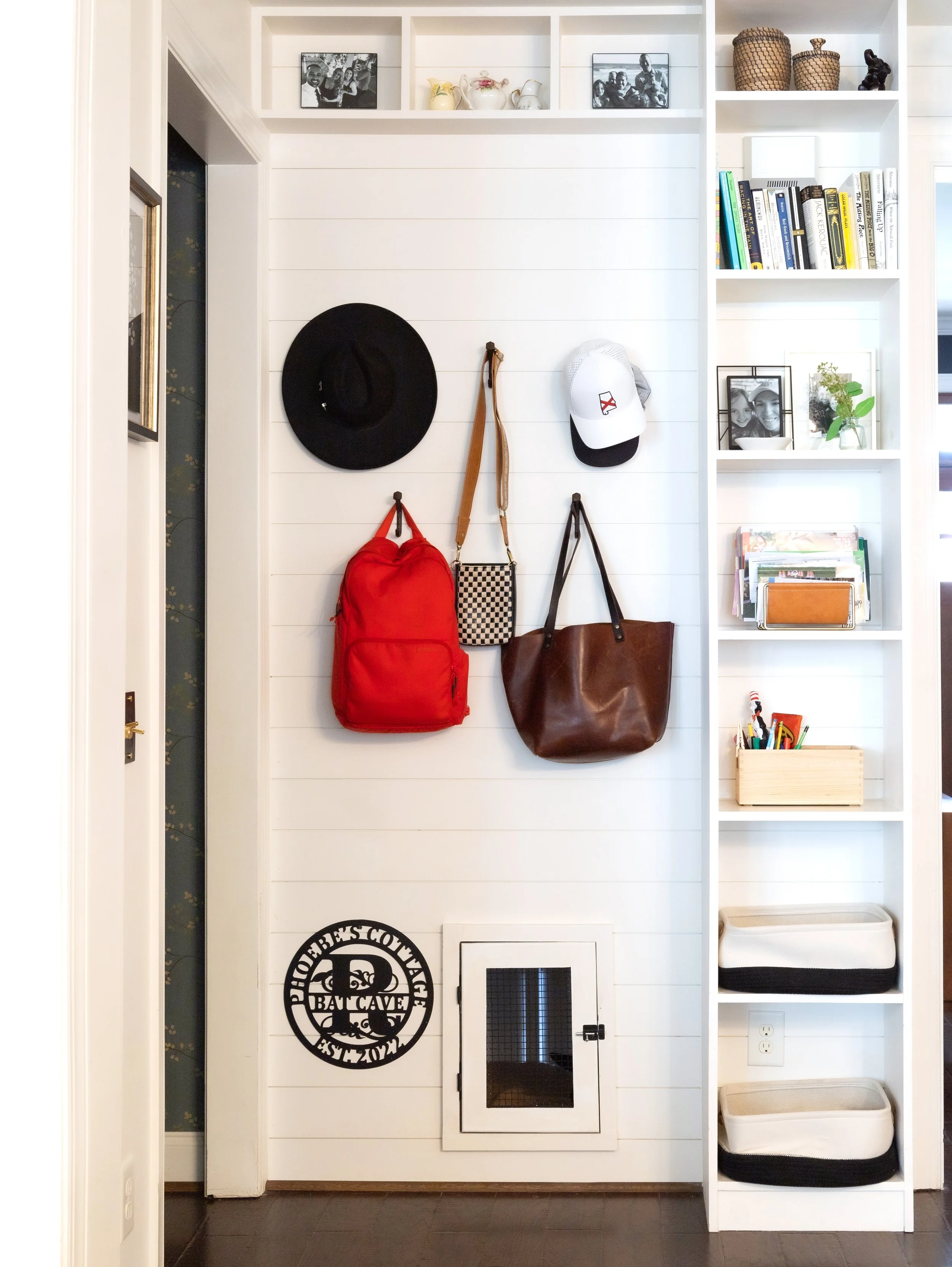 Entryway wall with hooks holding a black hat, white cap, red backpack, checkered bag, and brown leather tote. Top shelves hold black and white photos, decorative baskets, and books. Bottom shelves contain storage boxes and a wooden tray with art supplies. A small door in the wall is also visible.