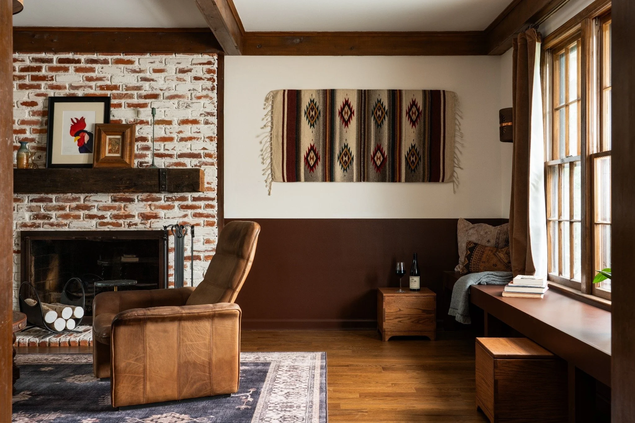 A cozy living room with a brick fireplace, a wood mantel decorated with framed art and small vases, leather chairs, a colorful Southwestern-style wall hanging, large windows with brown curtains, and wooden flooring.