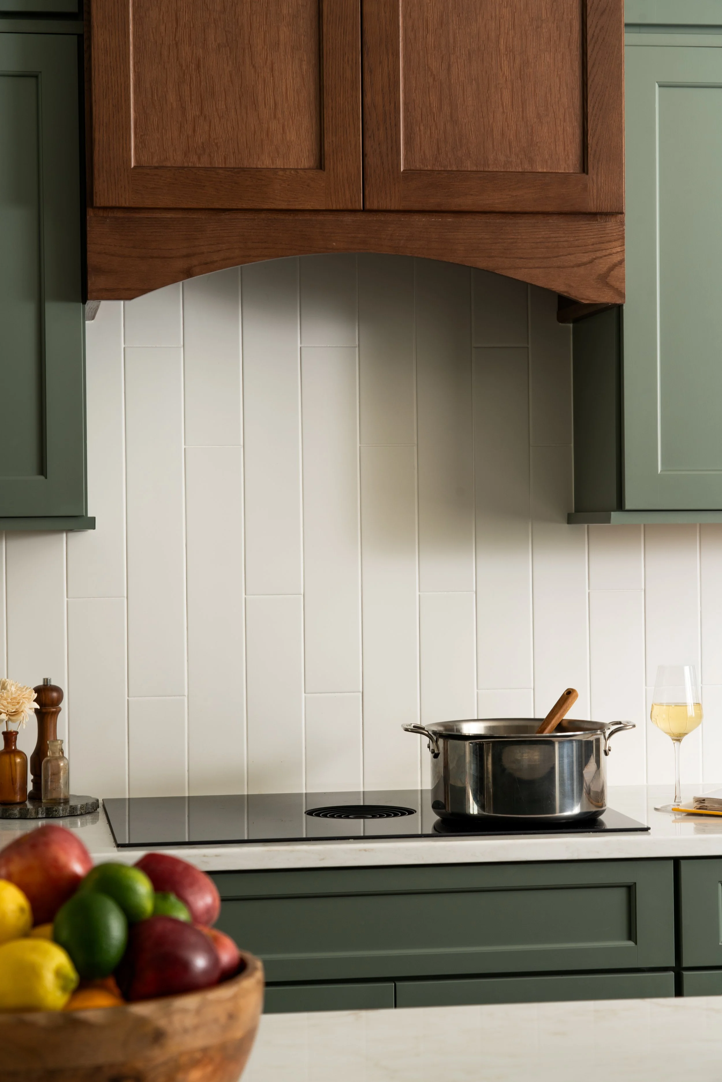 A kitchen with green cabinets, a wooden range hood, white tiled backsplash, and a stainless steel pot on the stove. There is a half-filled glass of white wine and a wooden spoon on the counter. A bowl of colorful apples and lemons is in the foregroun