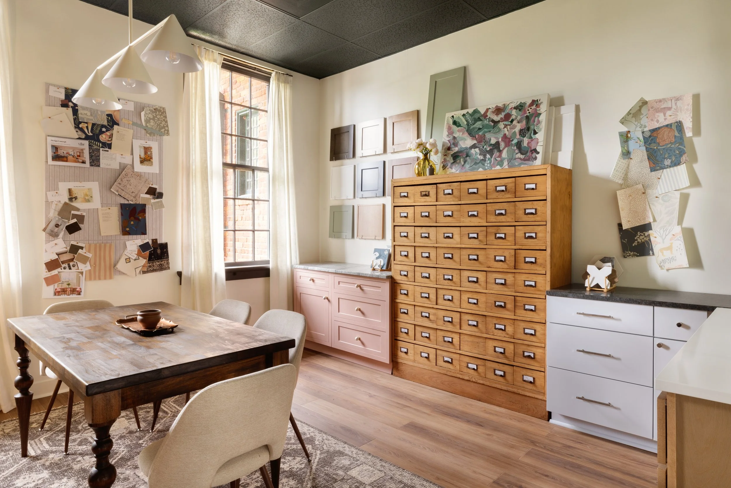 A cozy room with a wooden dining table and six beige upholstered chairs, a wooden chest of drawers on the wall decorated with art pieces and a yellow vase with flowers, a wall with picture samples, and a large window with curtains allowing natural light.
