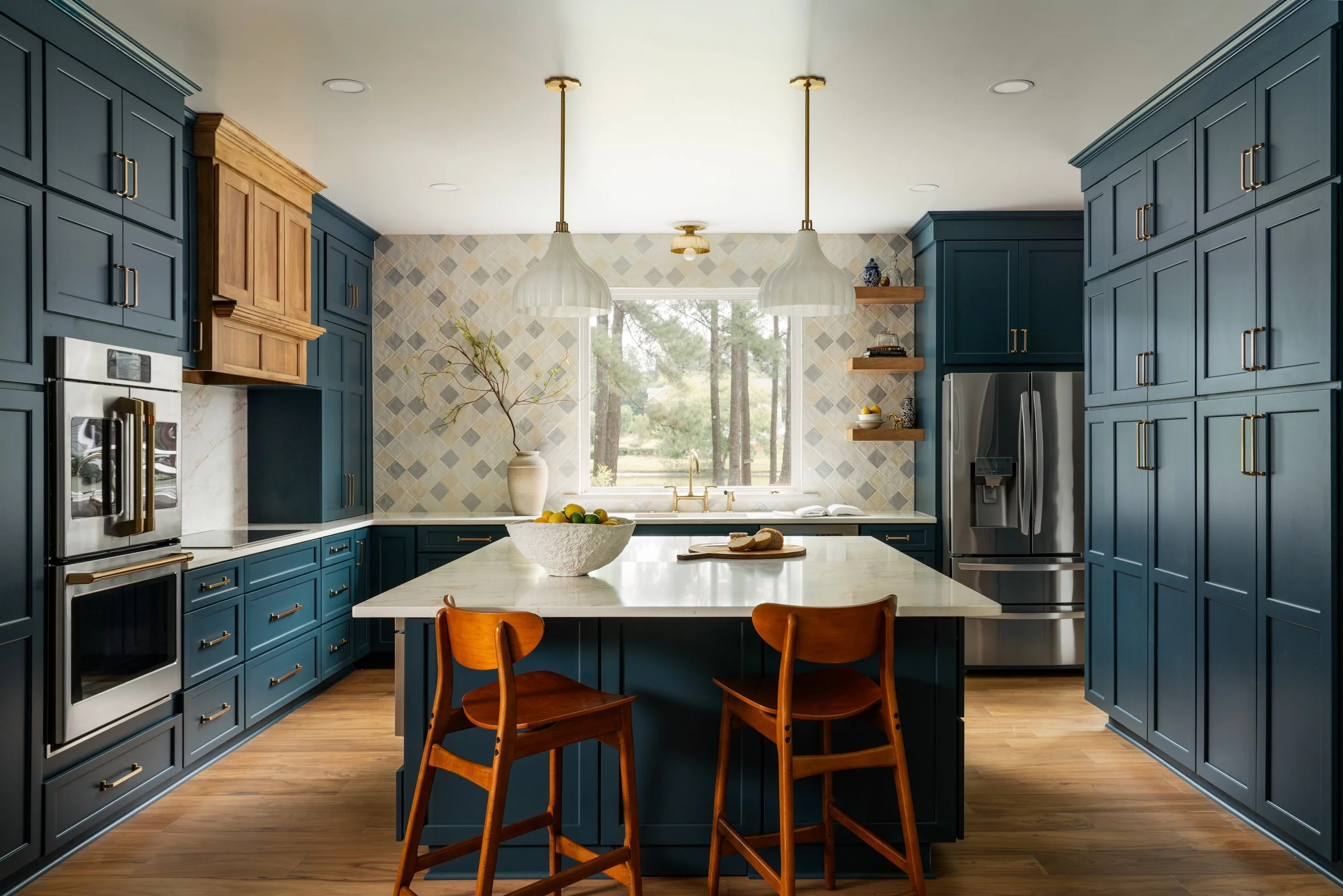A modern kitchen with blue cabinets, a white marble island, two pendant lights, and a window overlooking trees.
