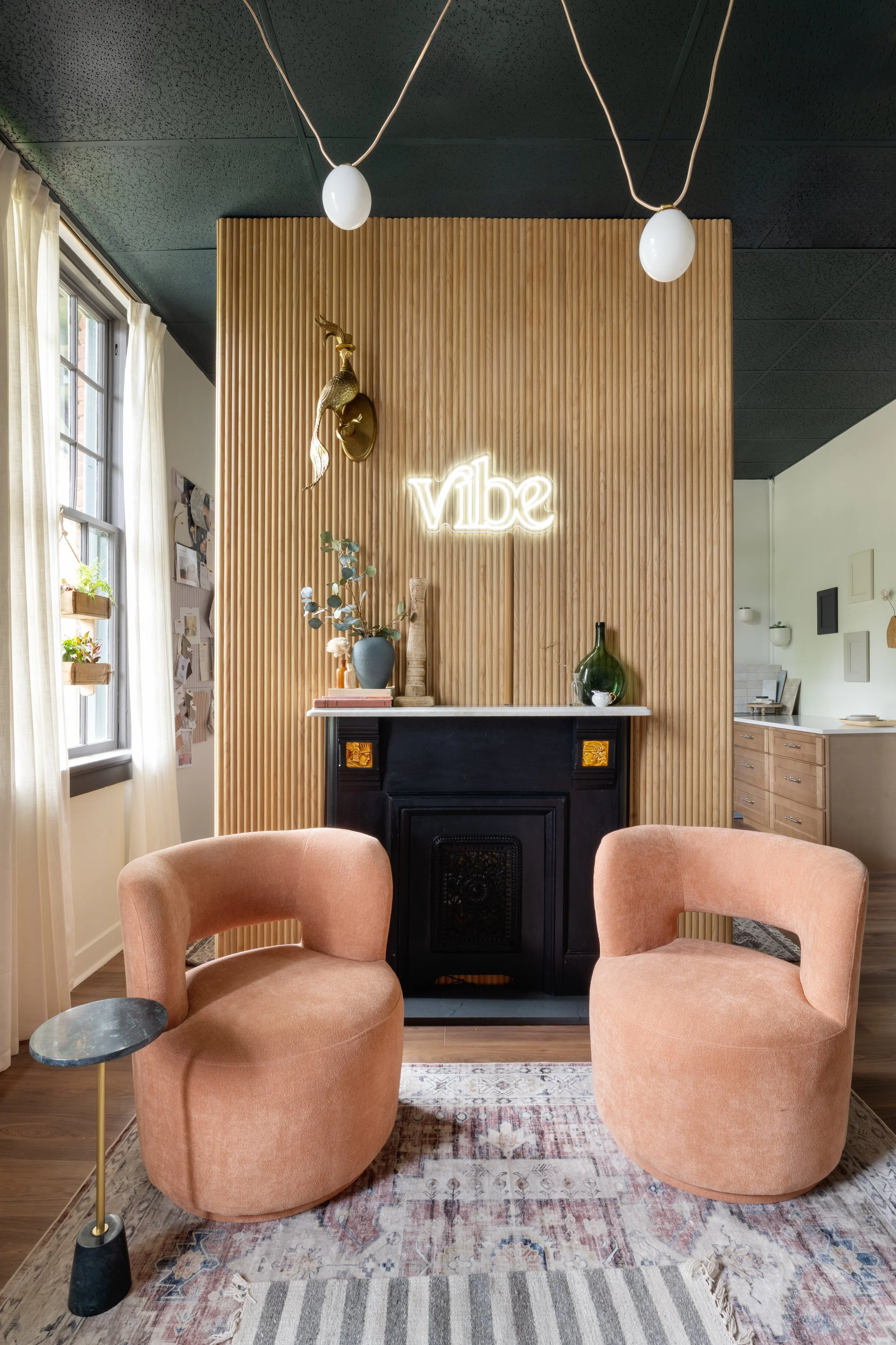 A cozy living room corner featuring two blush pink armchairs with rounded backs, a small side table with a dark marble top and gold base, a multicolored rug on wooden flooring, a decorative fireplace with a black grate, a wooden wall with vertical slats, a gold deer head wall sculpture, a neon sign that says 'vibe', a tall green glass vase, a blue ceramic vase with eucalyptus, a beige book, and a large window with white curtains.