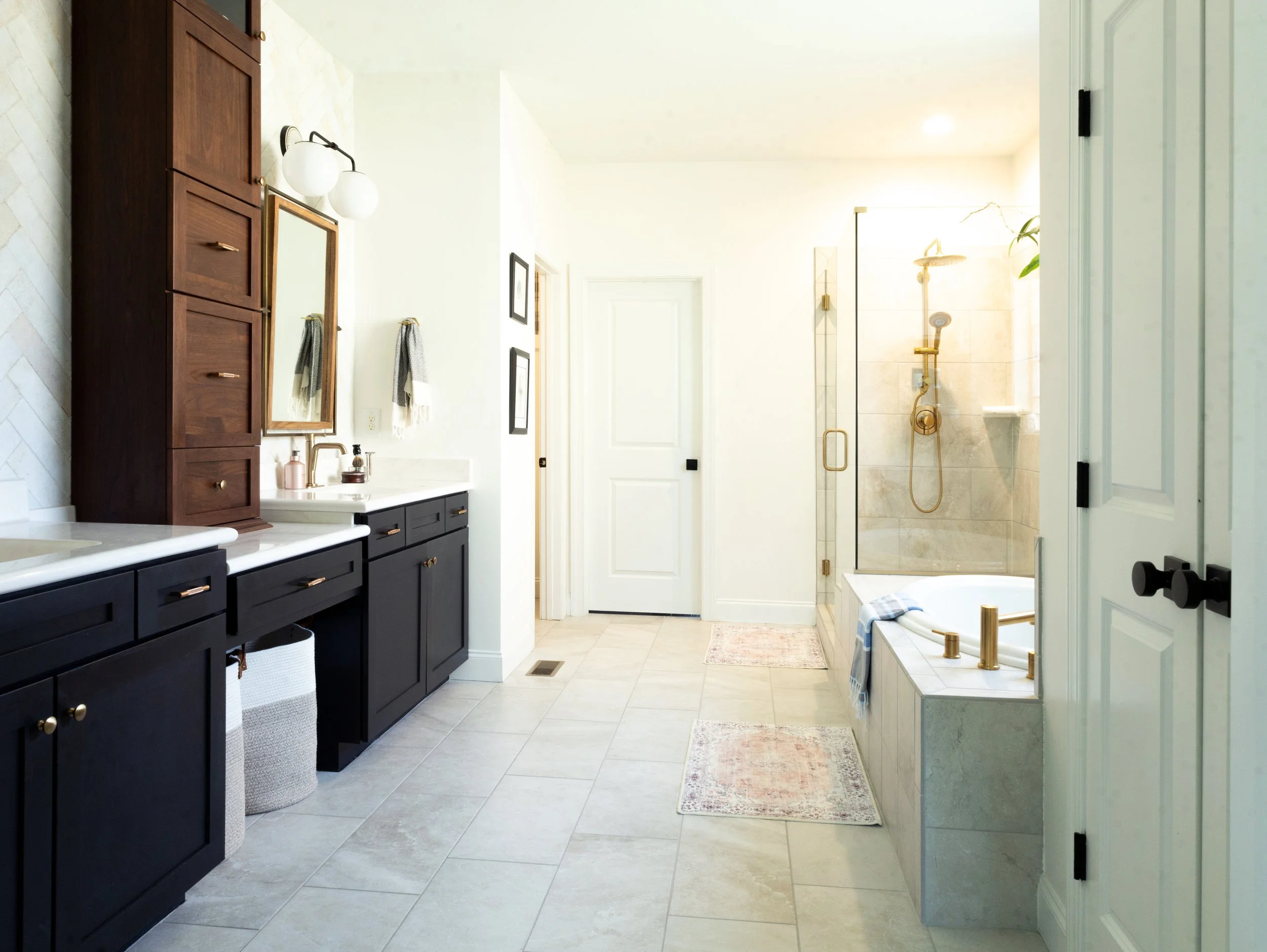 Bright bathroom with black and wooden cabinets, a double sink, towels, framed artwork, a shower with glass door and gold fixtures, and a built-in bathtub with gold fixtures.