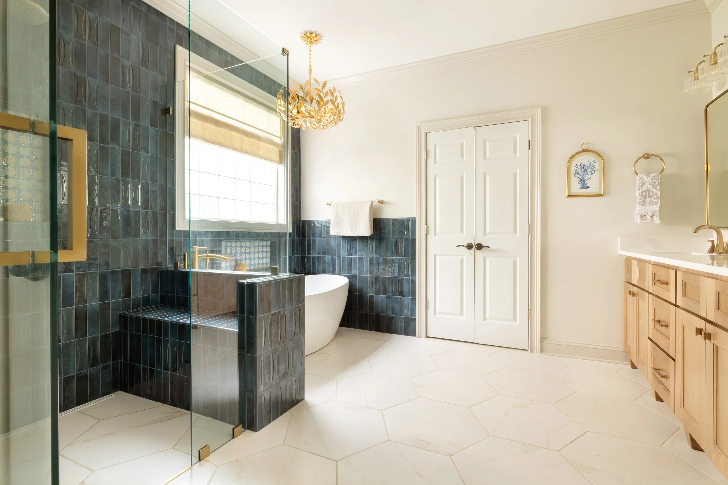 A bathroom with a walk-in glass shower with gold fixtures, dark blue tiled walls, a white freestanding bathtub, a window with a beige roman shade, a white door, a towel rack, framed blue coral artwork, and a wooden vanity with a white countertop and 