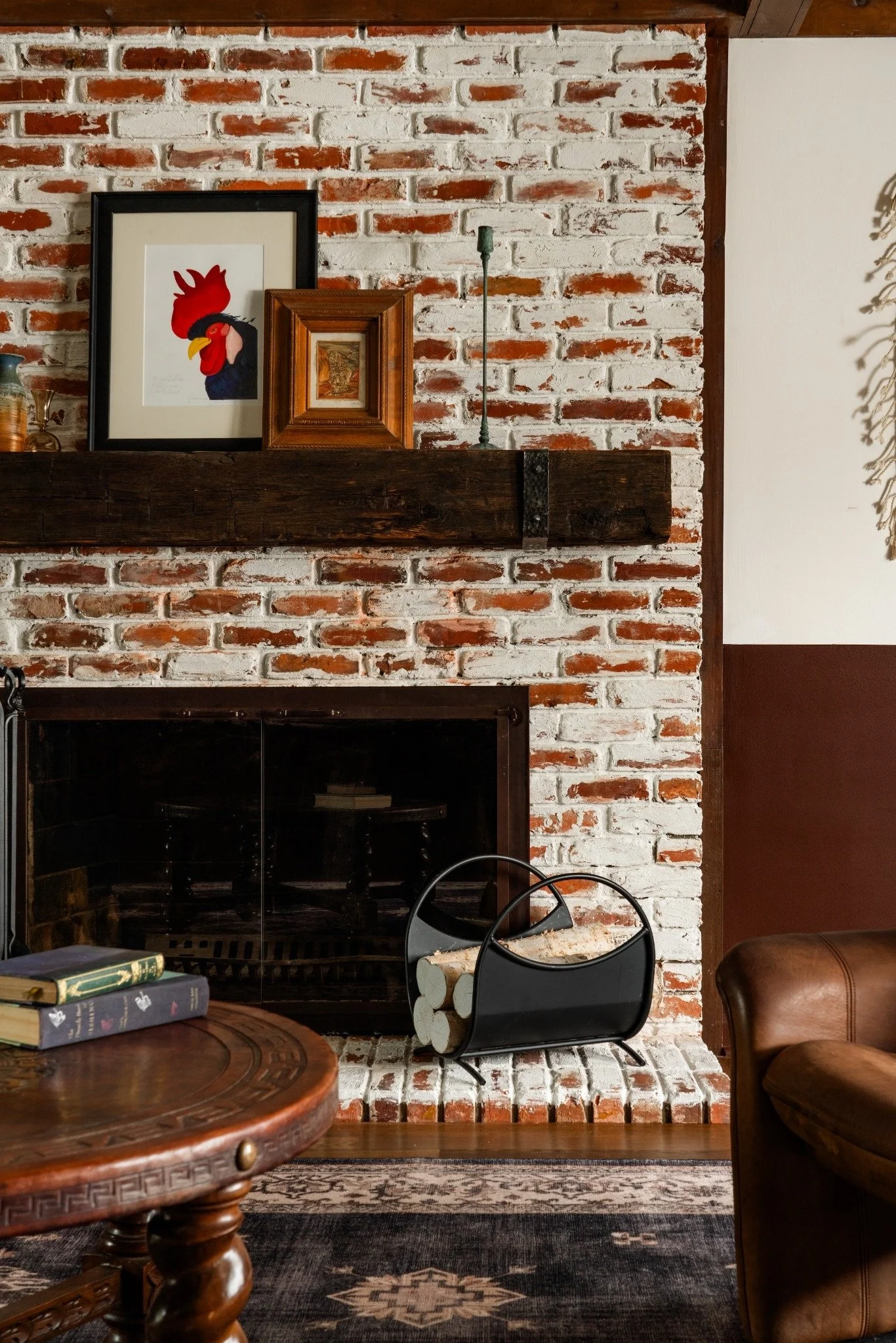 A brick fireplace with a wooden mantle holding framed artwork, a brass candlestick, and decorative logs. Below the mantle, there's a black metal log holder, a leather armchair, a patterned rug, and a wooden table with books and a plant. To the right,