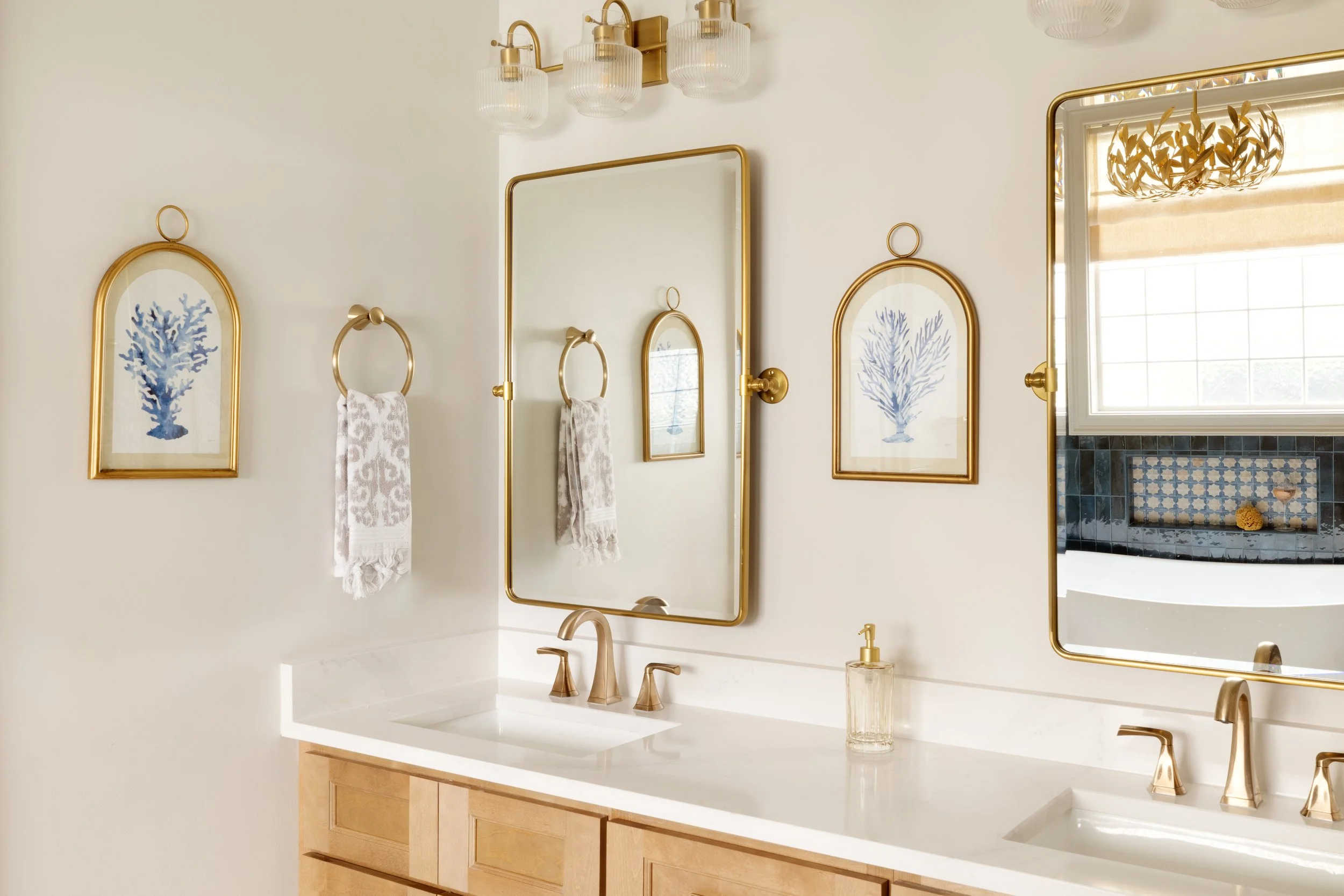 A bathroom with white walls, decorated with gold-framed pictures of blue coral, gold towel rings, and large mirrors. There are three wall-mounted light fixtures with ribbed glass shades above the mirrors. The bathroom has a white countertop with a go