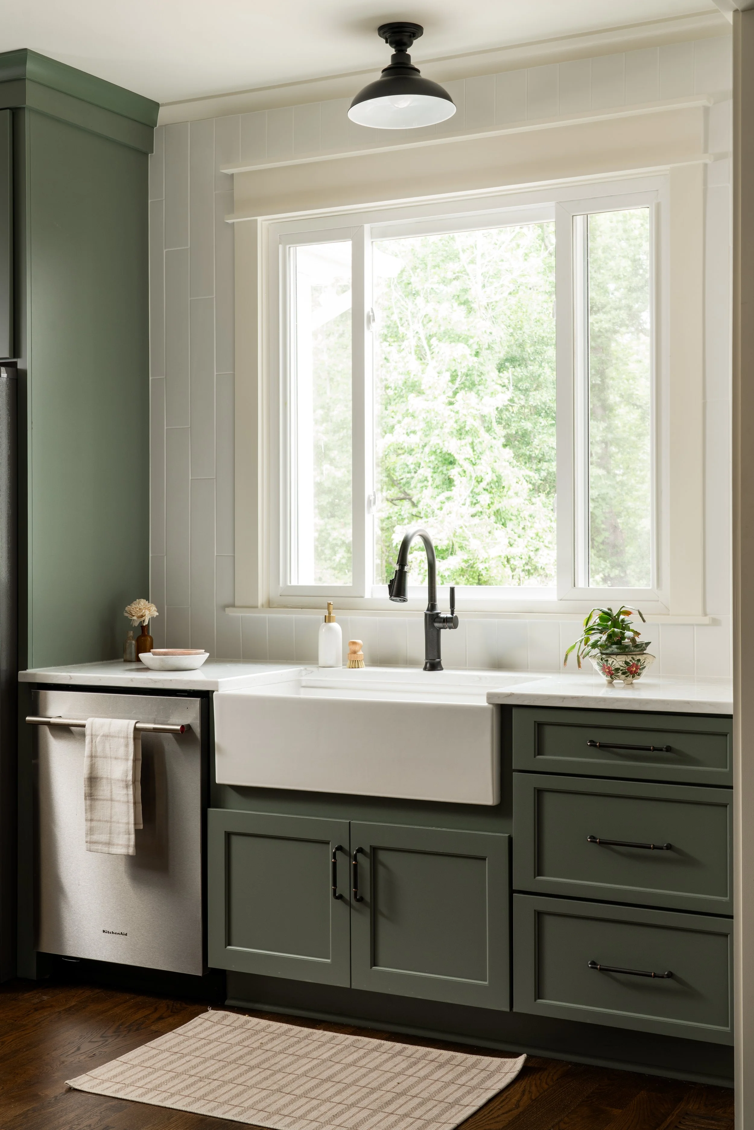 Kitchen with green cabinets, white farmhouse sink, and window looking out to greenery, decorated with potted plant, soap dispenser, and small vase with flowers, with dishwasher and beige rug.