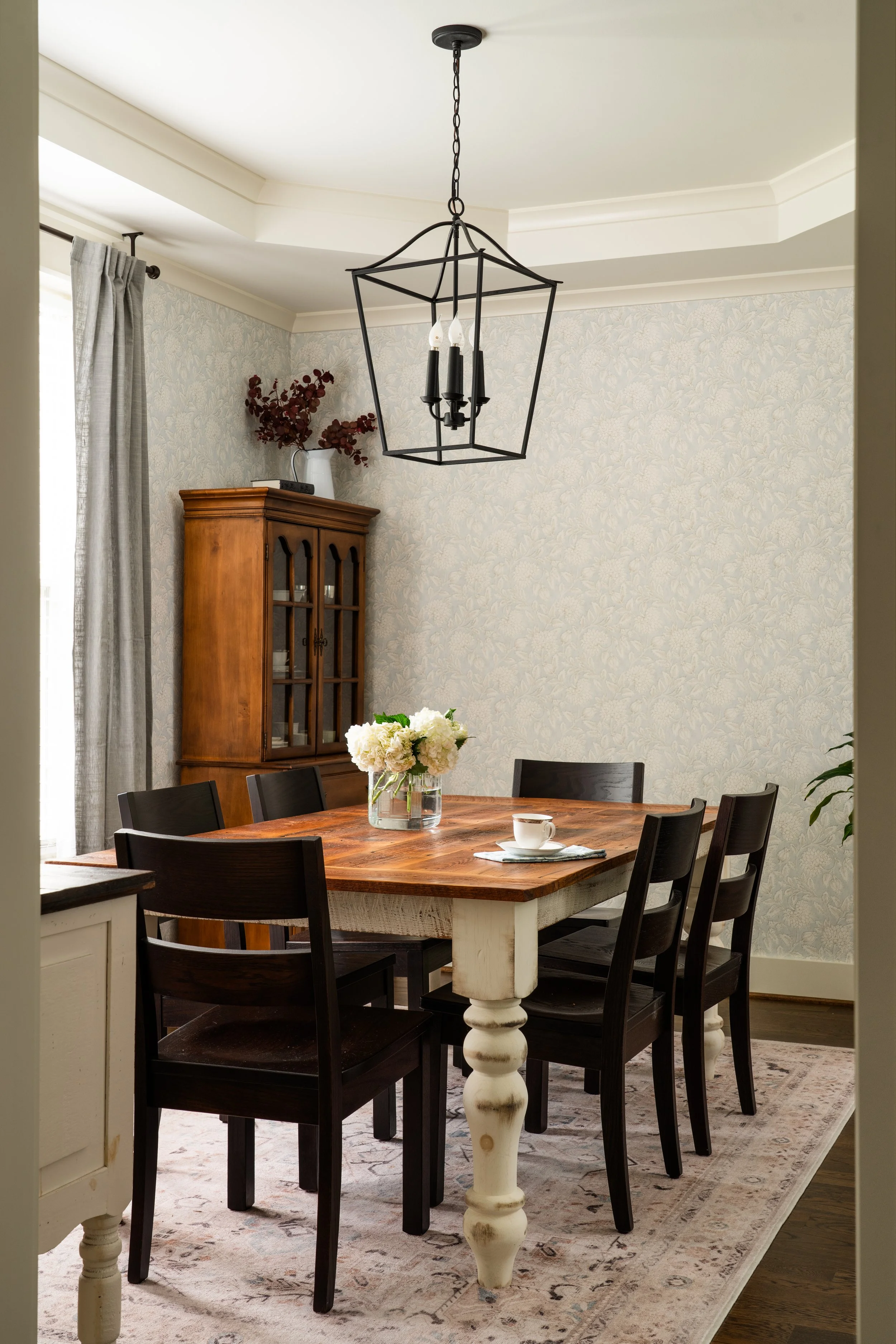 A dining room with a wooden table, six black chairs, a floral centerpiece, a rug, and a cabinet with glass doors. There's a chandelier hanging above the table and light-colored wallpaper on the walls.