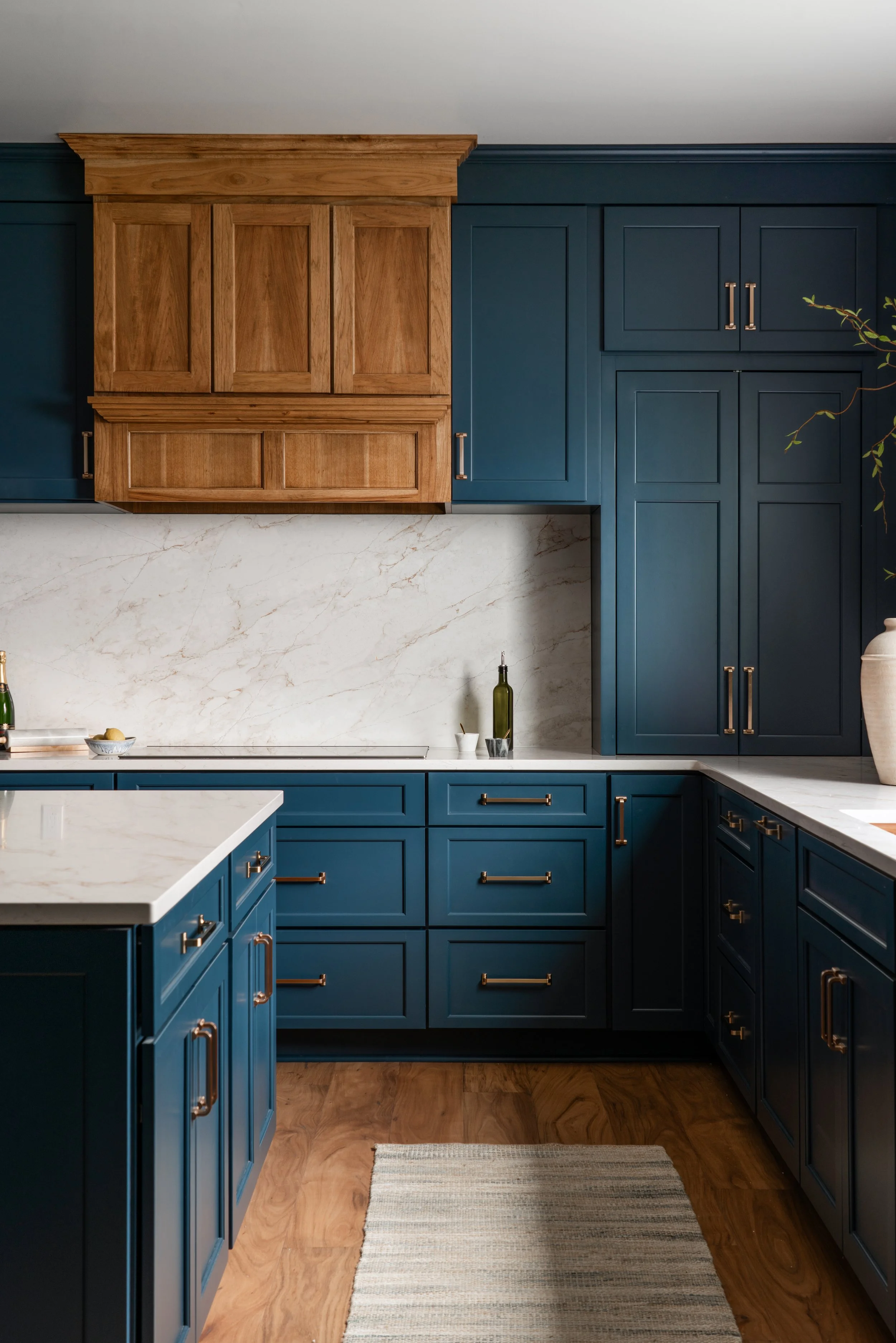 Kitchen with blue cabinets, wooden upper cabinets, marble backsplash, and wood flooring, with a white countertop and a striped rug on the floor.