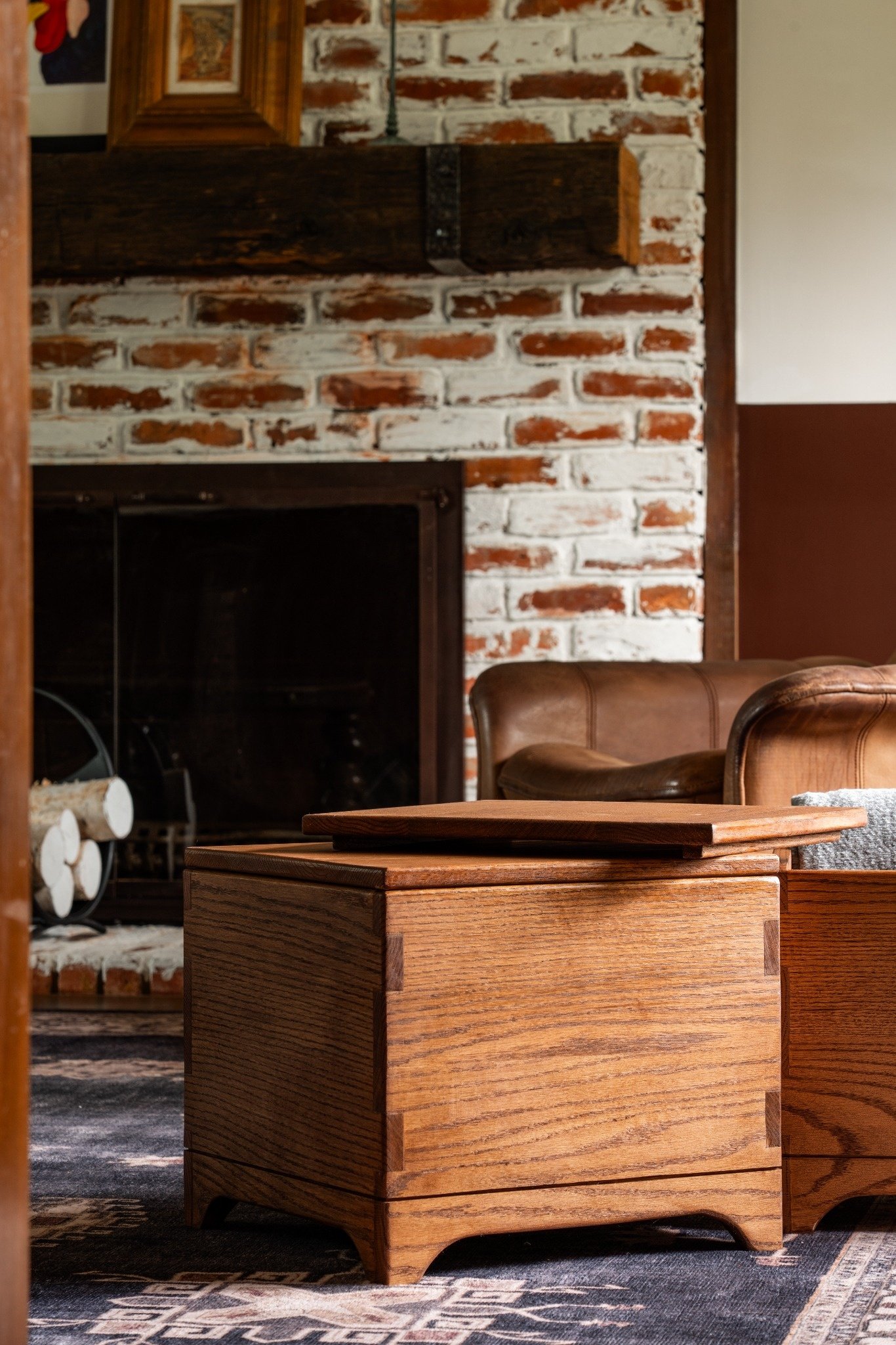 A close-up of a wooden box on a patterned rug in front of a brick fireplace, with leather chairs nearby in a cozy living room.