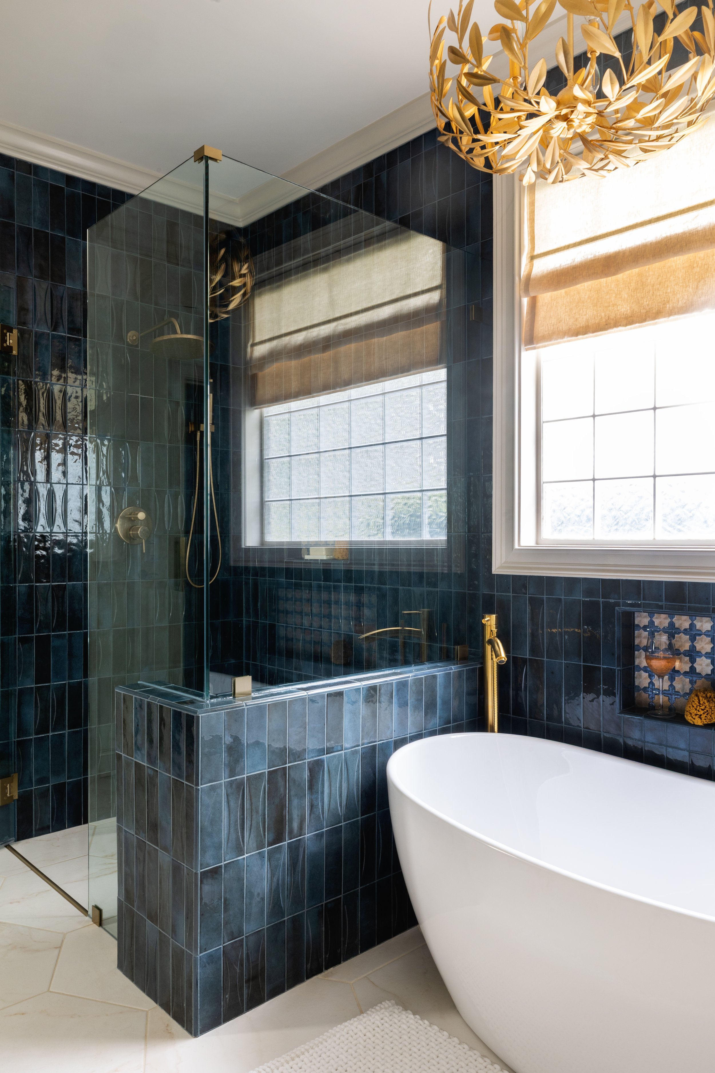 A bathroom with a white freestanding bathtub, a dark blue tiled shower area with a glass partition, and a large window with beige Roman shades. A gold chandelier hangs from the ceiling.