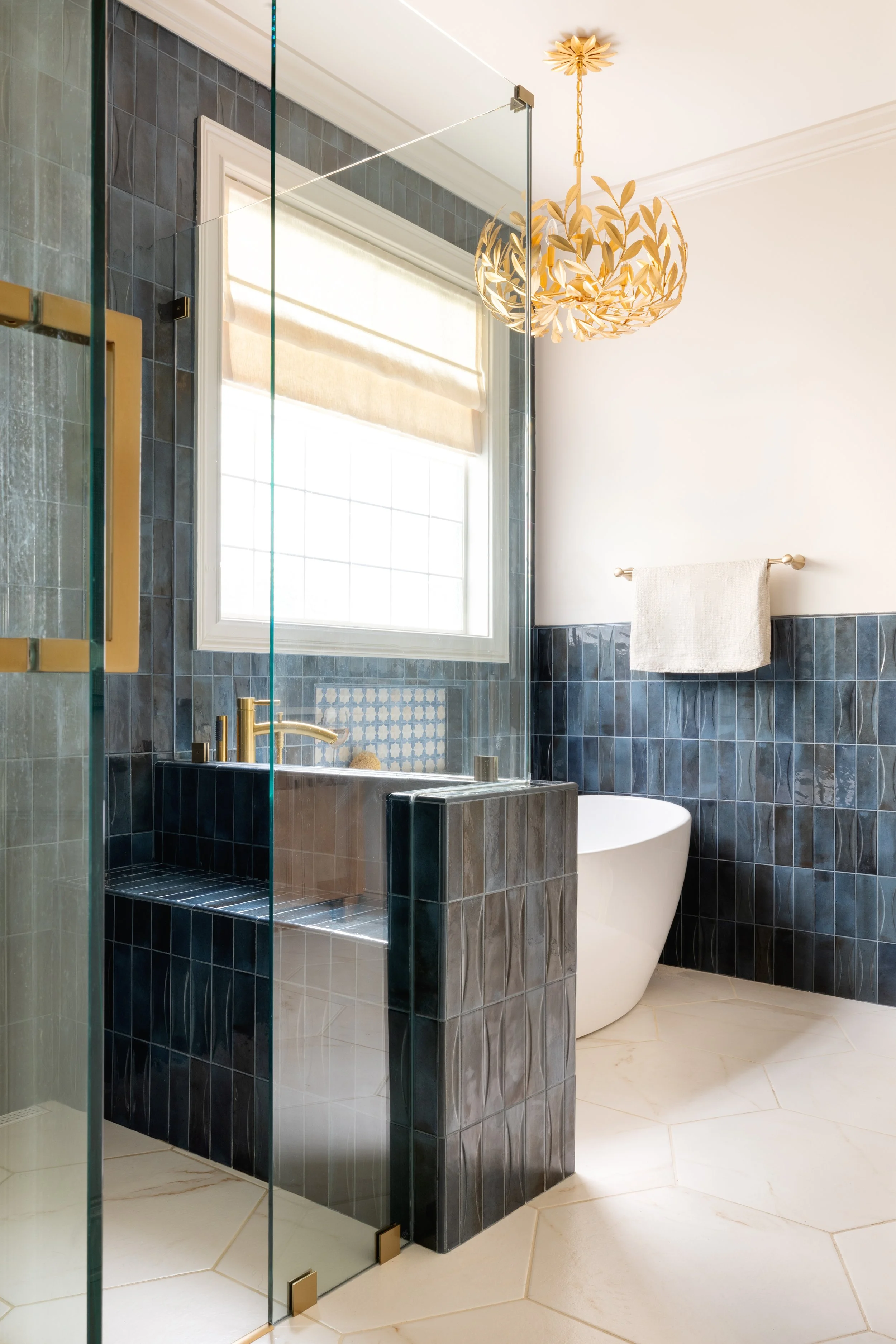 Modern bathroom with a blue tile wall, a white window with blinds, a gold chandelier, a towel rack with a towel, and a walk-in shower with glass doors and a gold handle.