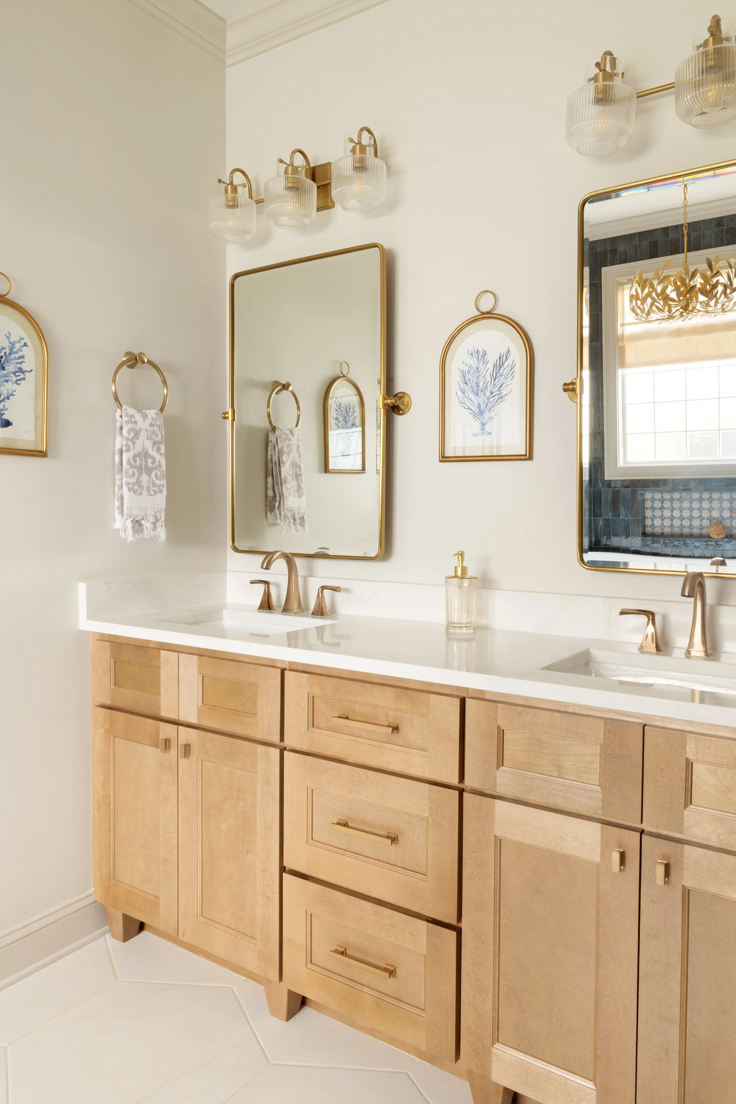 A bathroom vanity with a white countertop, two sinks, gold fixtures, a large mirror, framed art featuring a blue coral, two wall-mounted towel rings with patterned towels, and trimmed black and white tiles on the walls reflected in the mirror.