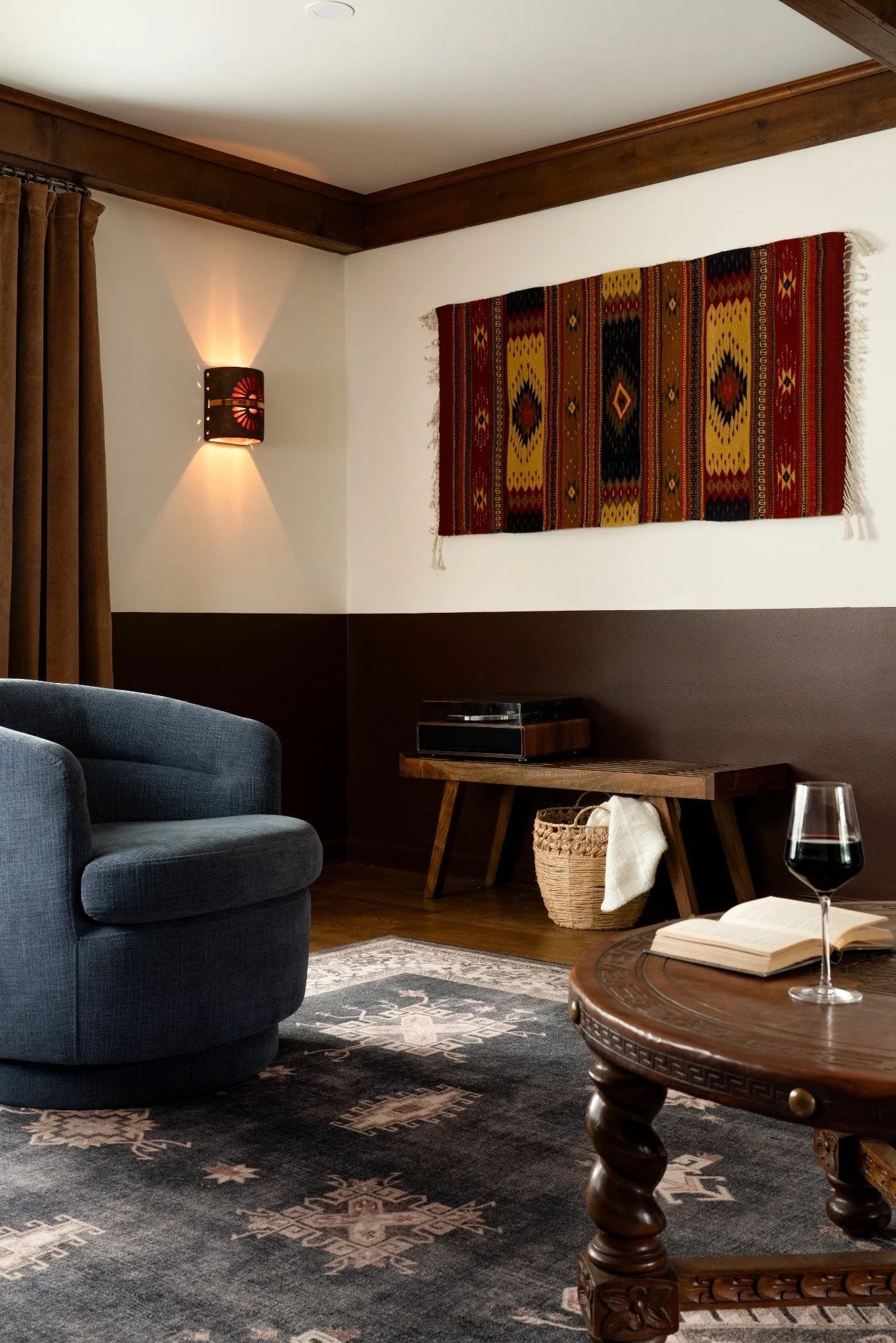 A cozy living room with a dark patterned rug, blue armchair, wooden table with a glass of red wine and open book, wooden side table with a record player, wall with a colorful woven wall hanging, wall sconce, and brown curtains.