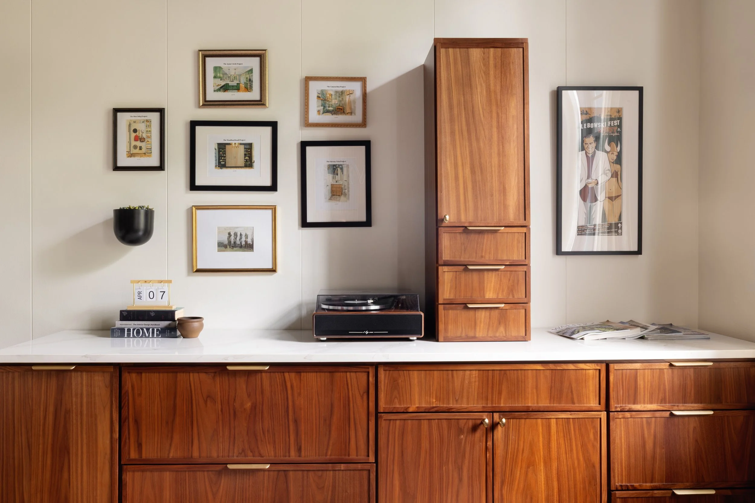A wooden kitchen cabinet with a built-in bookshelf, framed pictures on the wall, a potted plant, a calendar, and a stack of magazines on a white countertop, with a small stereo or speaker below the framed pictures.