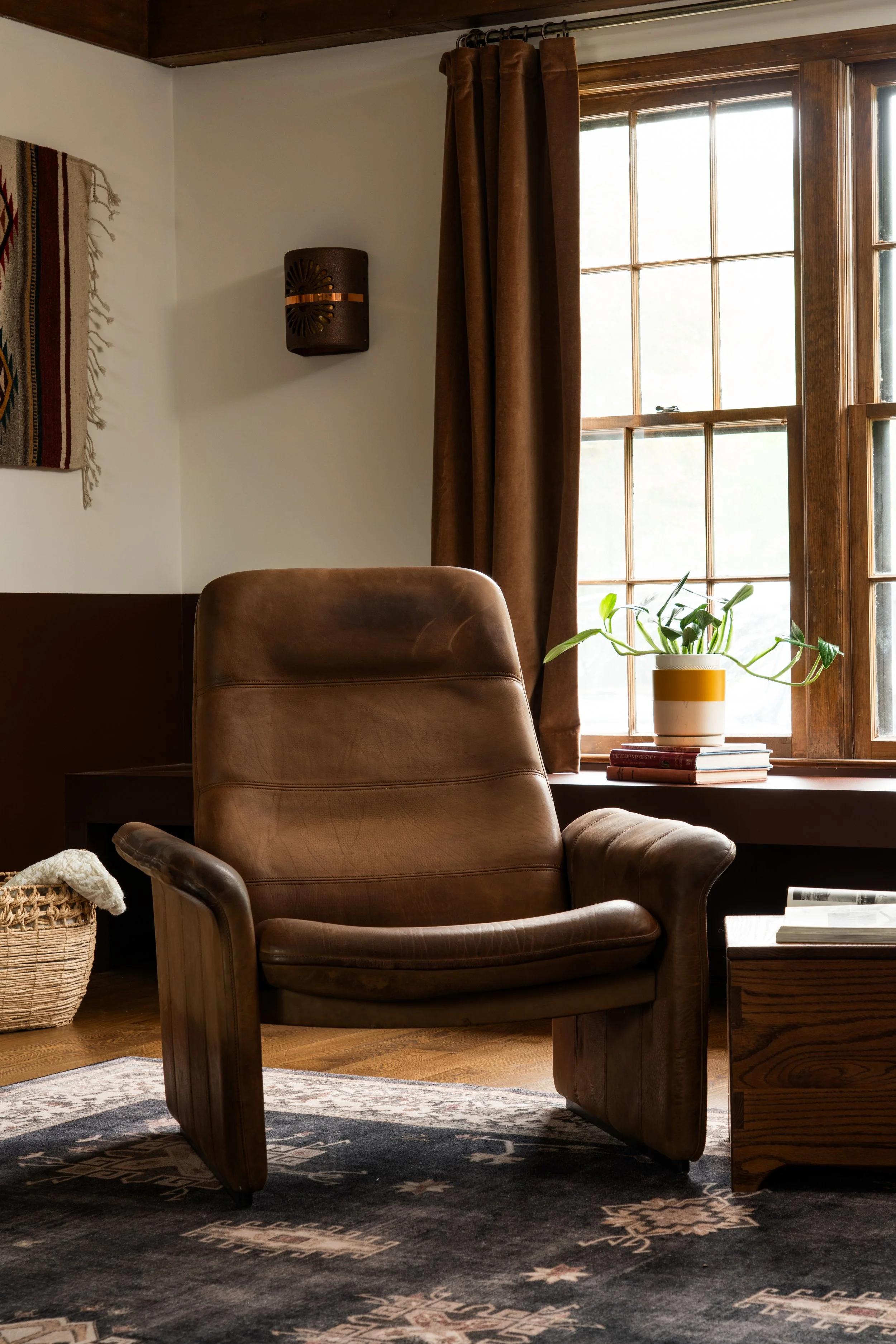 A cozy reading corner featuring a leather armchair, a side table with books, a potted plant on top, a window with wooden trim and brown curtains, a woven basket on the floor, a wall hanging, and a decorative dark wall sconce. Natural light streams in