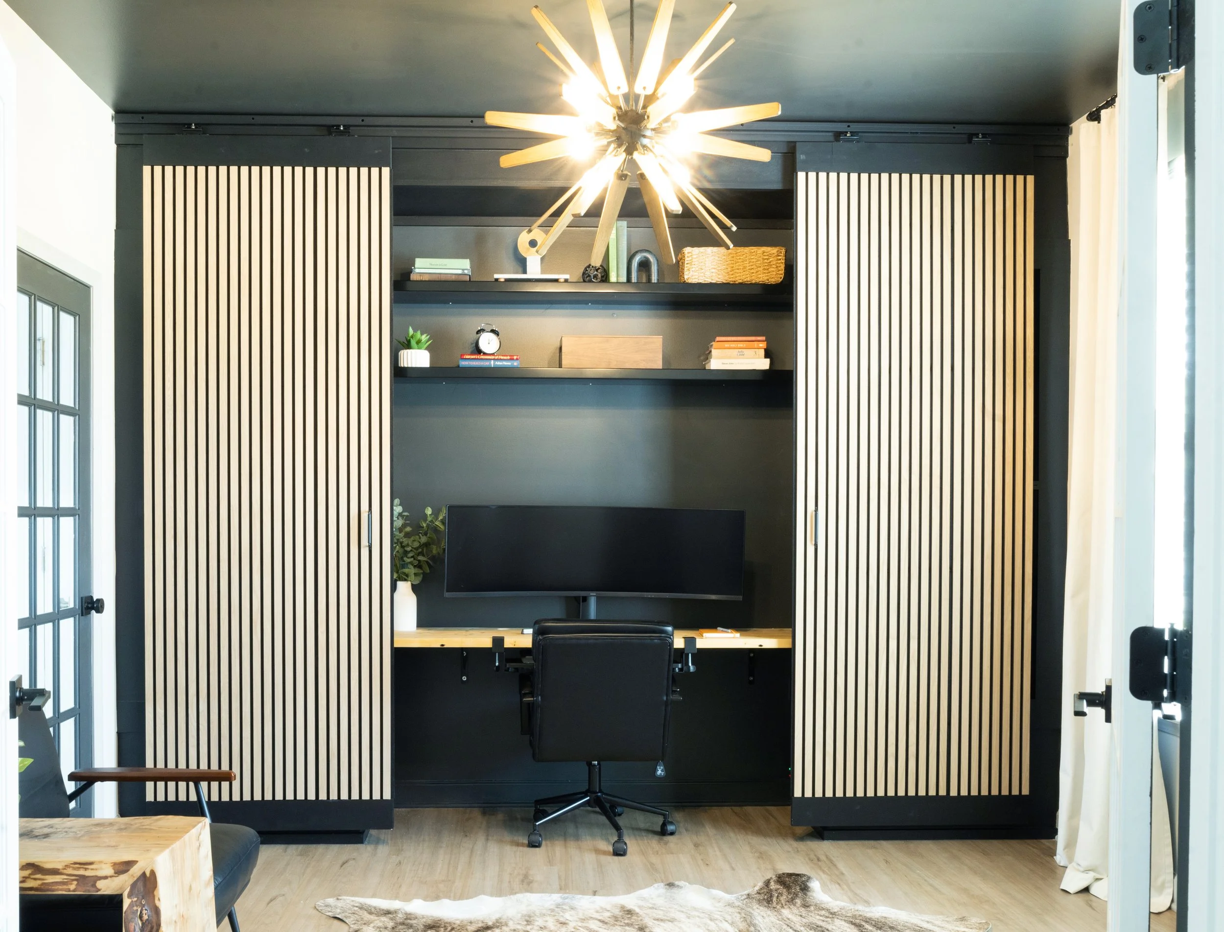 Modern home office with black wall, built-in shelving, a large curtain on the right, a desk with a computer monitor and chair, and a unique wooden atom-inspired ceiling light fixture.