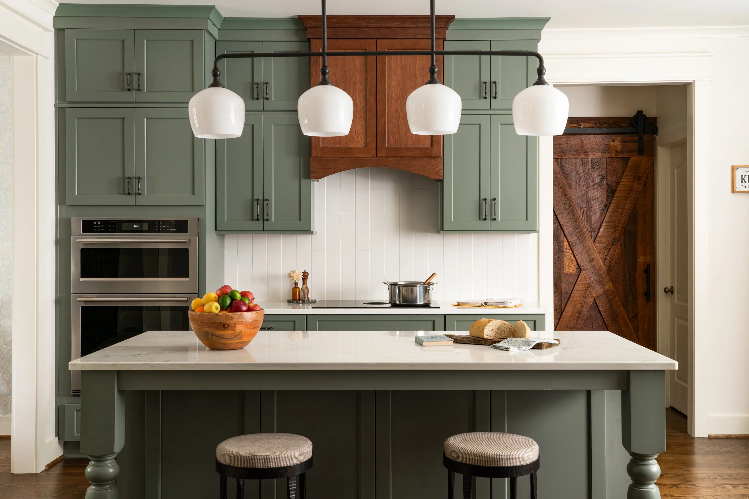 Kitchen with green cabinets, a white marble island, a wooden bowl of apples, a loaf of bread, stools, and a wooden sliding barn door.
