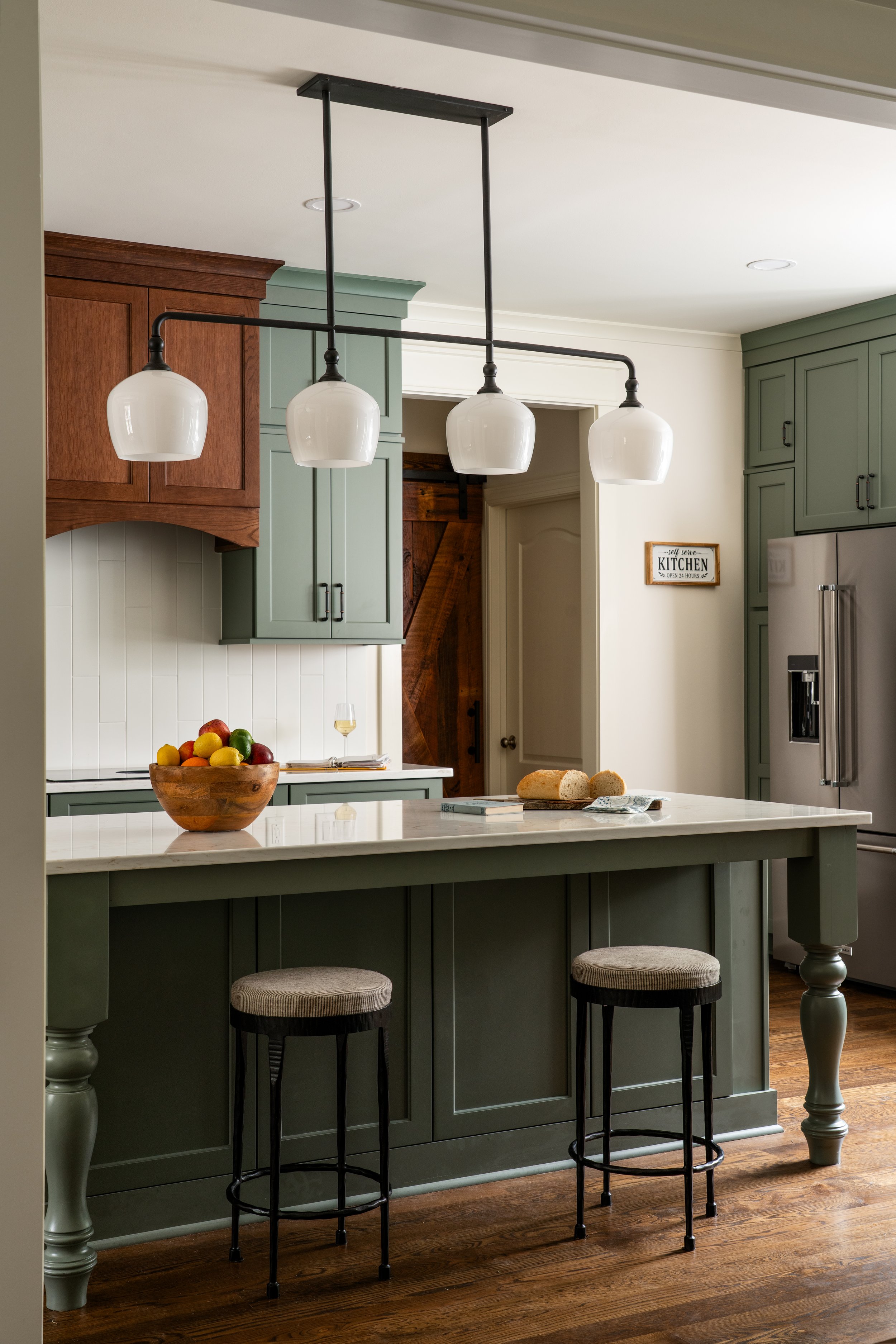 A modern kitchen with green cabinets, a white island, a bowl of colorful fruit, and three white pendant lights hanging above the island.
