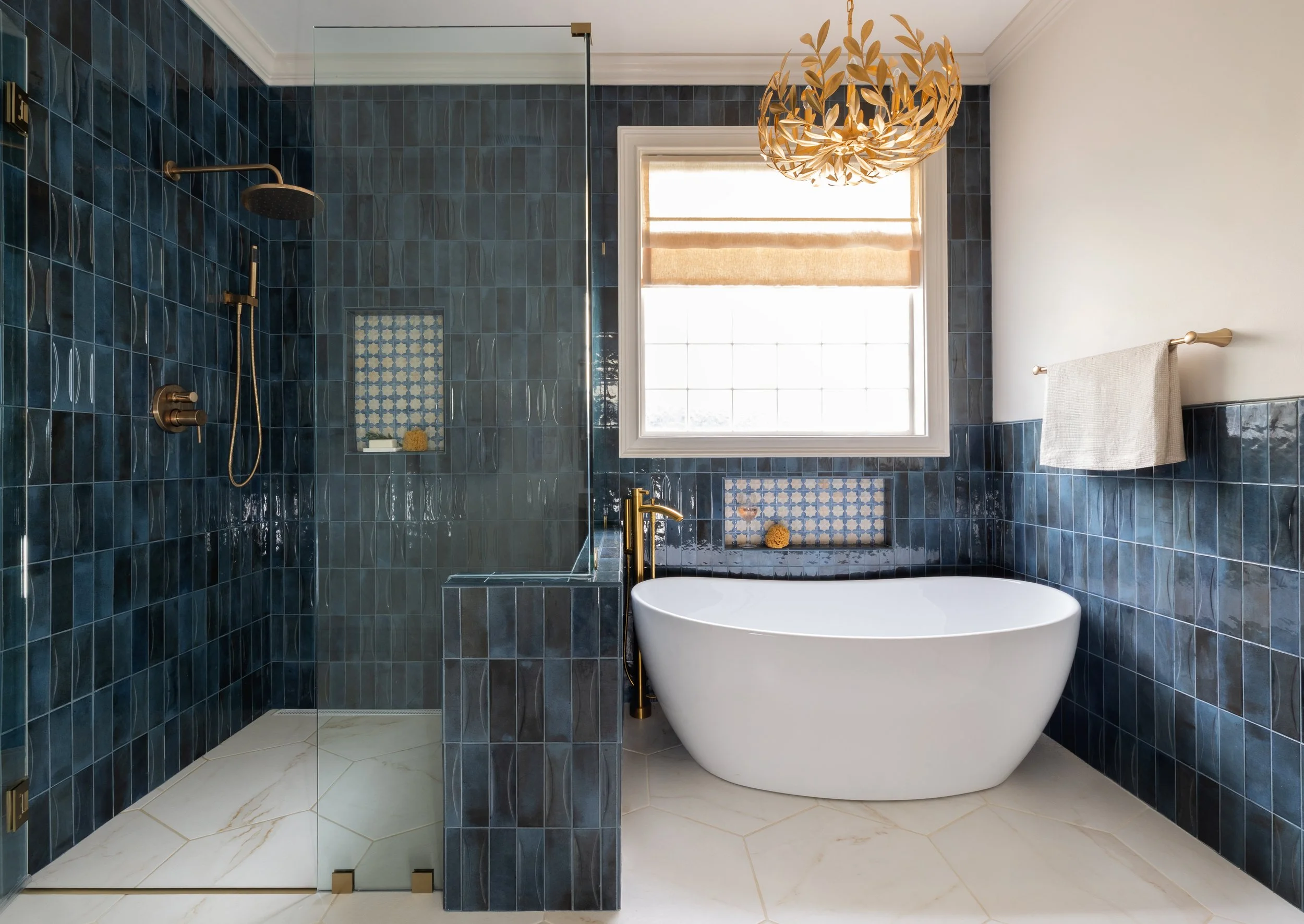 A modern bathroom featuring a blue tile shower area with a glass door and a built-in niche, a freestanding white bathtub, a window with a beige Roman shade, a beige towel on a rod, and a decorative pendant light fixture.