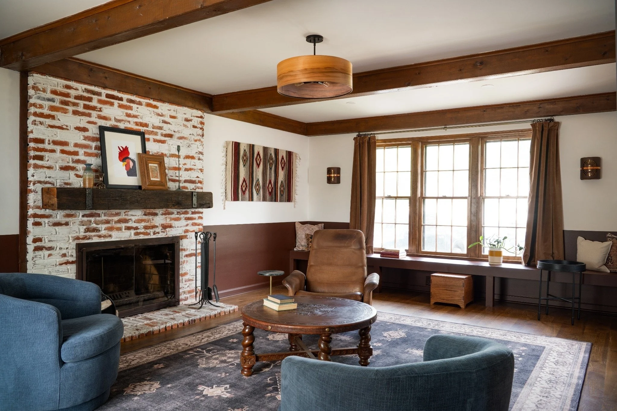 Living room with brick fireplace, wooden beams ceiling, large window with brown curtains, and vintage furniture including a leather armchair, blue upholstered chair, and wooden coffee table.