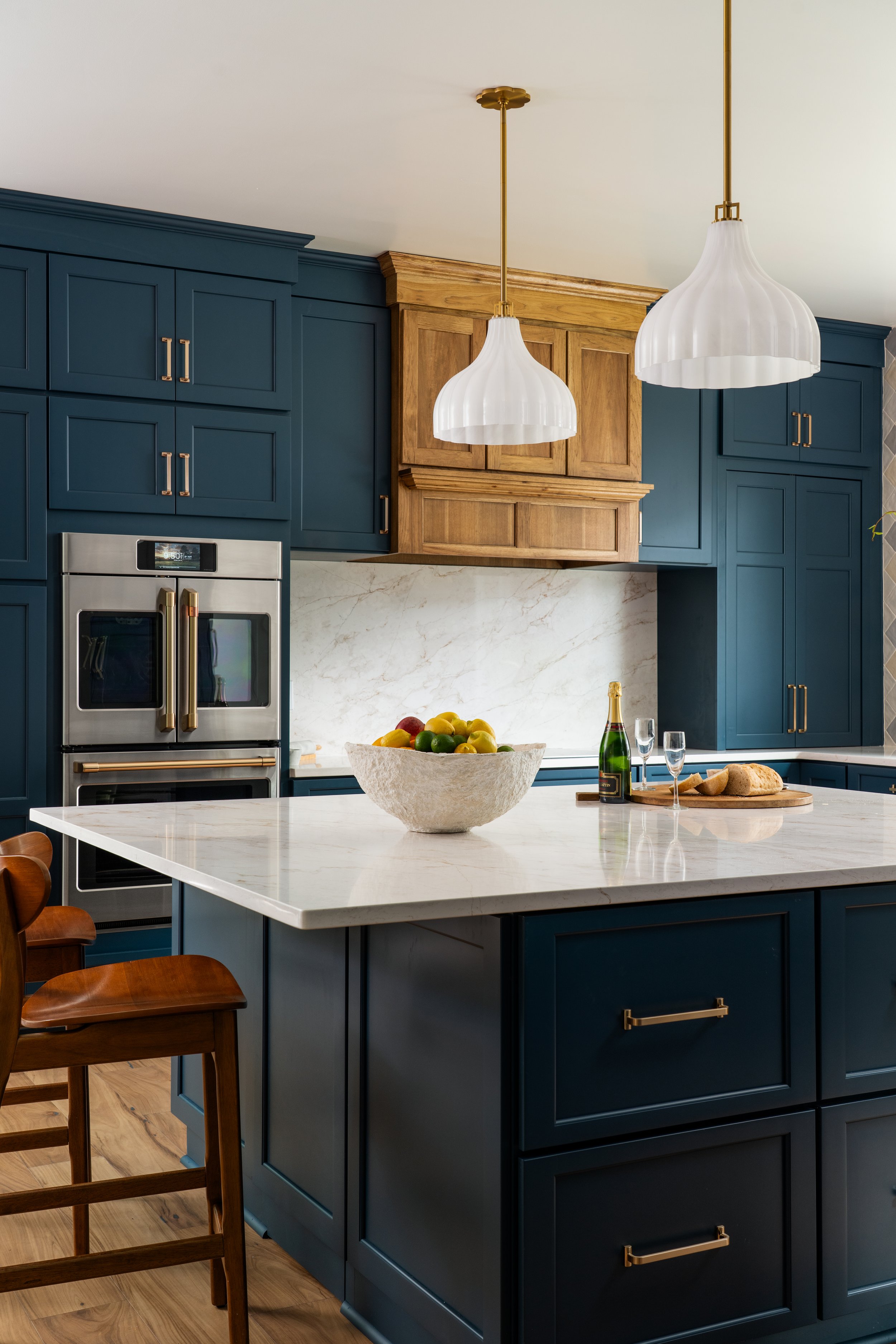 A kitchen with navy blue cabinets, a white marble countertop, and two white pendant lights hanging from a ceiling. There are wooden chairs around a kitchen island, a bowl of mixed fruit, a bottle of champagne, and two champagne glasses on the counter