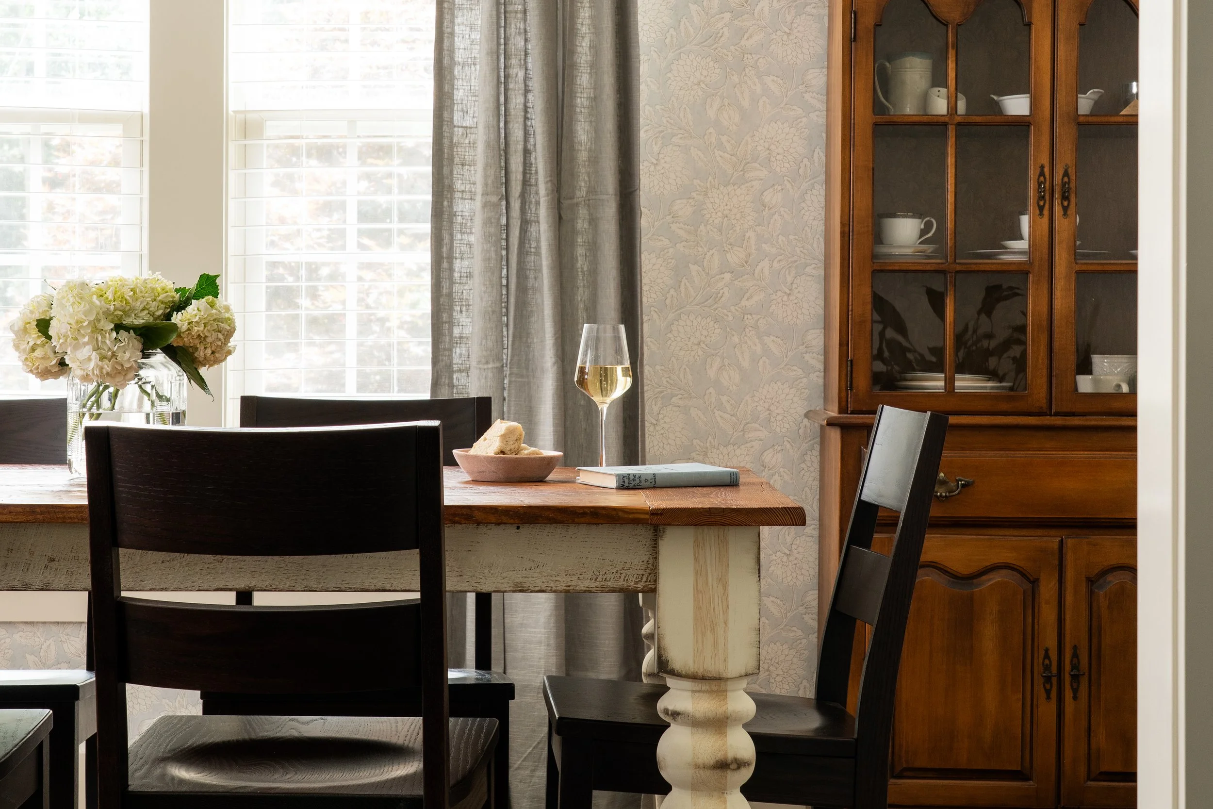 A dining room with a wooden table, black chairs, a vase of white hydrangeas, a glass of white wine, a bowl with bread, a magazine, a window with white blinds, a gray curtain, and a wooden China cabinet filled with dishware.