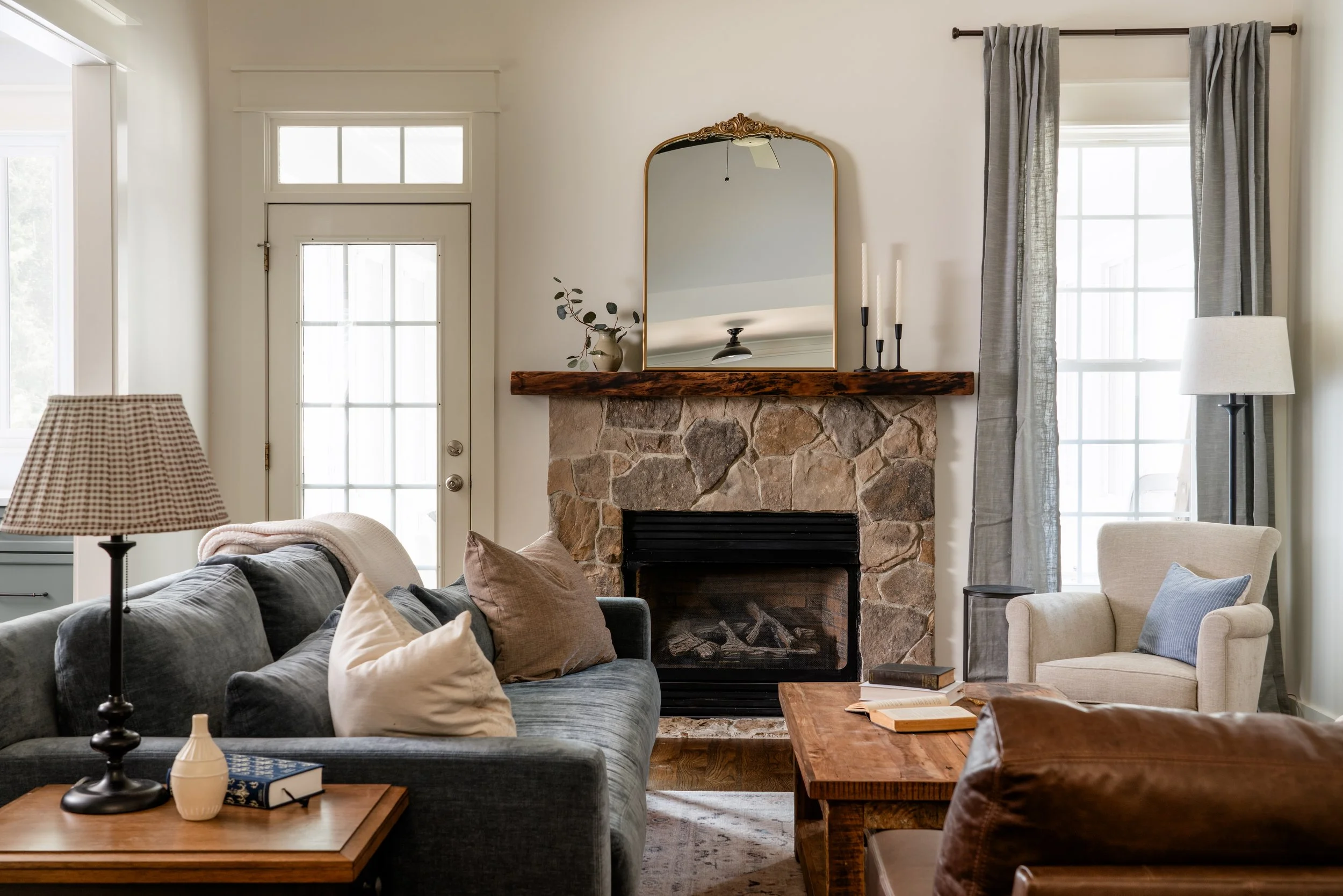 Cozy living room with a stone fireplace, mirror above mantel, beige and gray furniture, window with gray curtains, table lamp, and decorated coffee table.