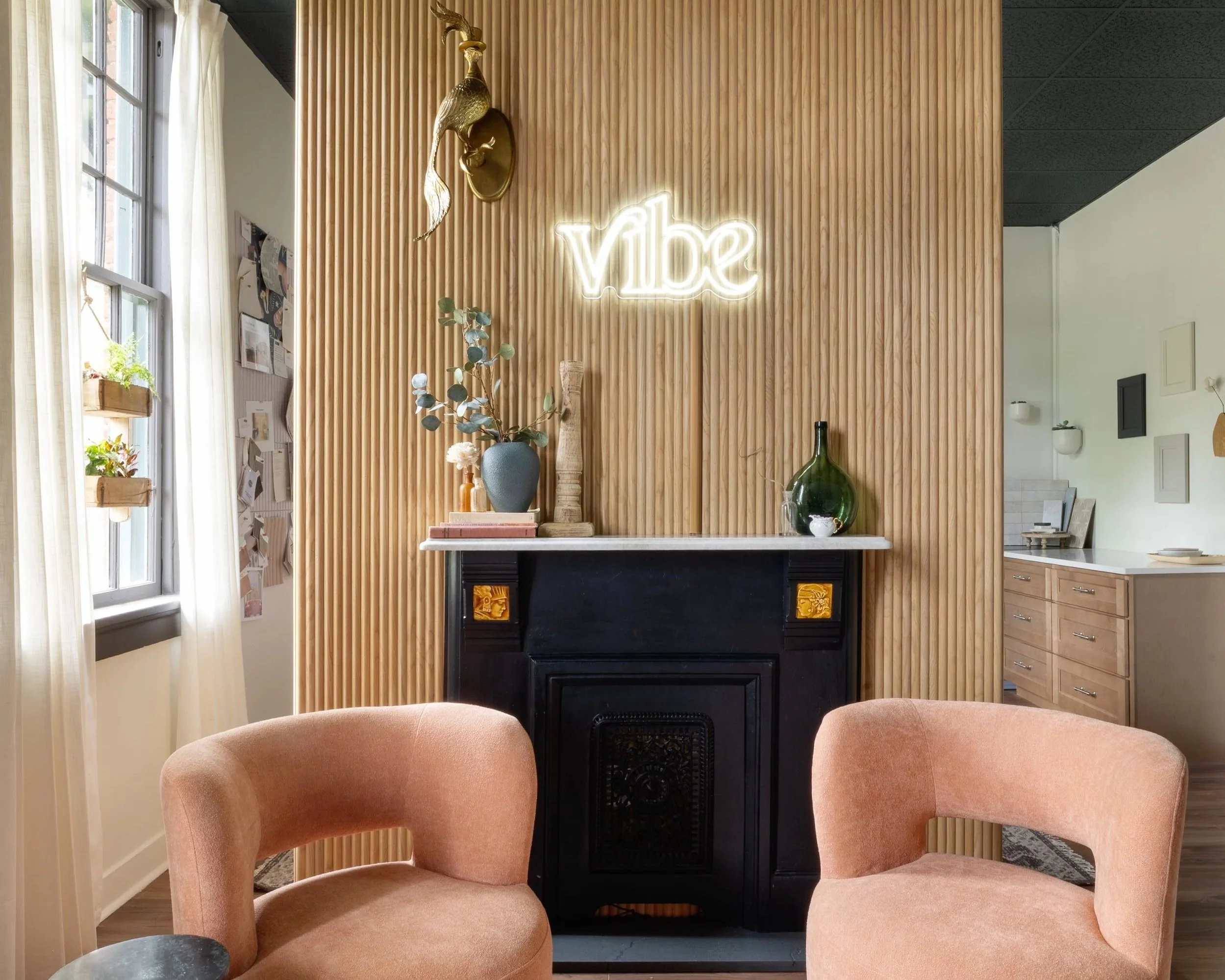 Interior of a room with a wooden slat wall, a neon sign that says "Vibe", a decorative bird sculpture, a vase with eucalyptus leaves on a table, and peach-colored upholstered chairs.
