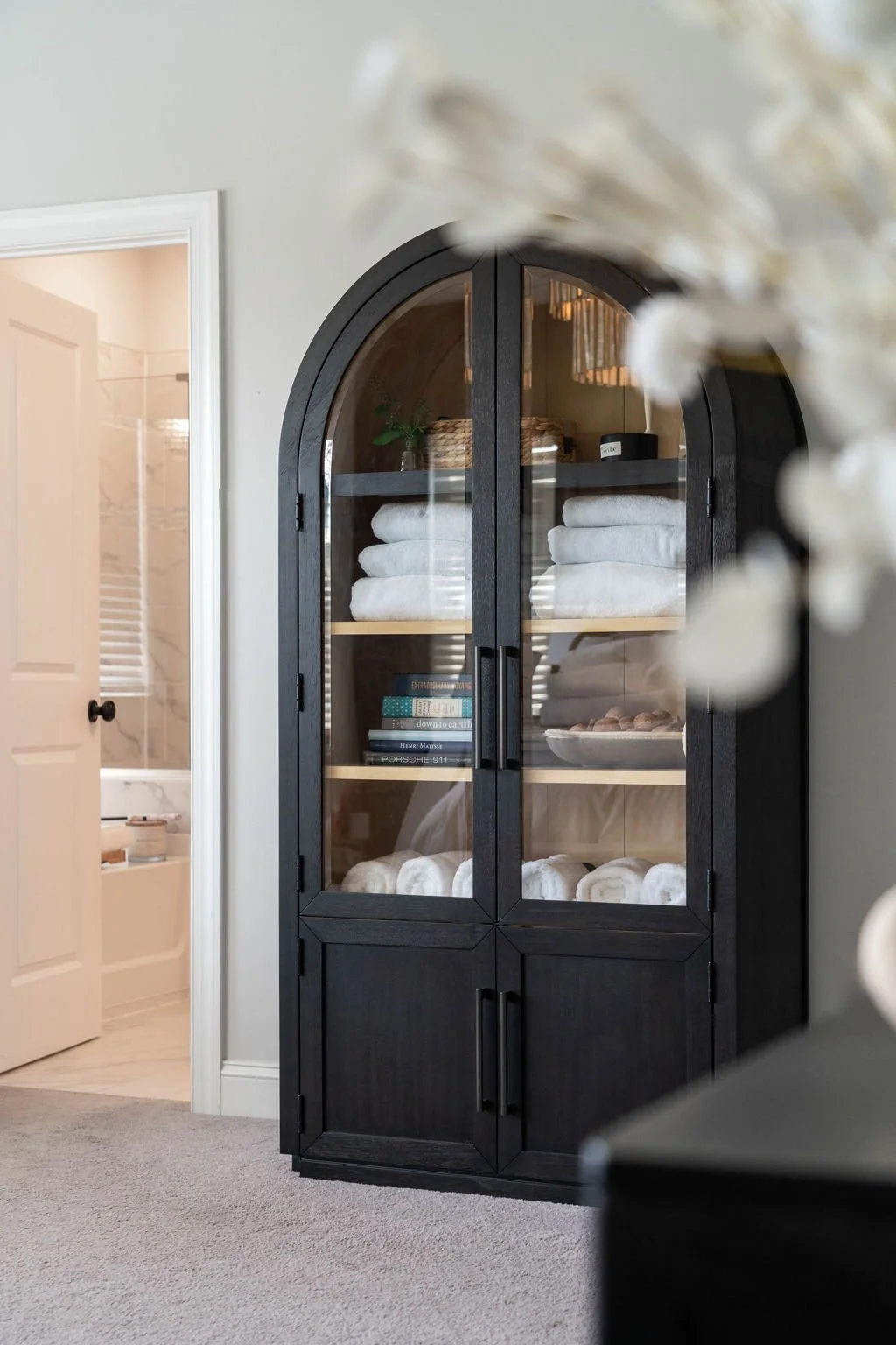 Black arched glass-front cabinet filled with neatly folded white towels, books, and decorative items, in a modern bathroom or bedroom.