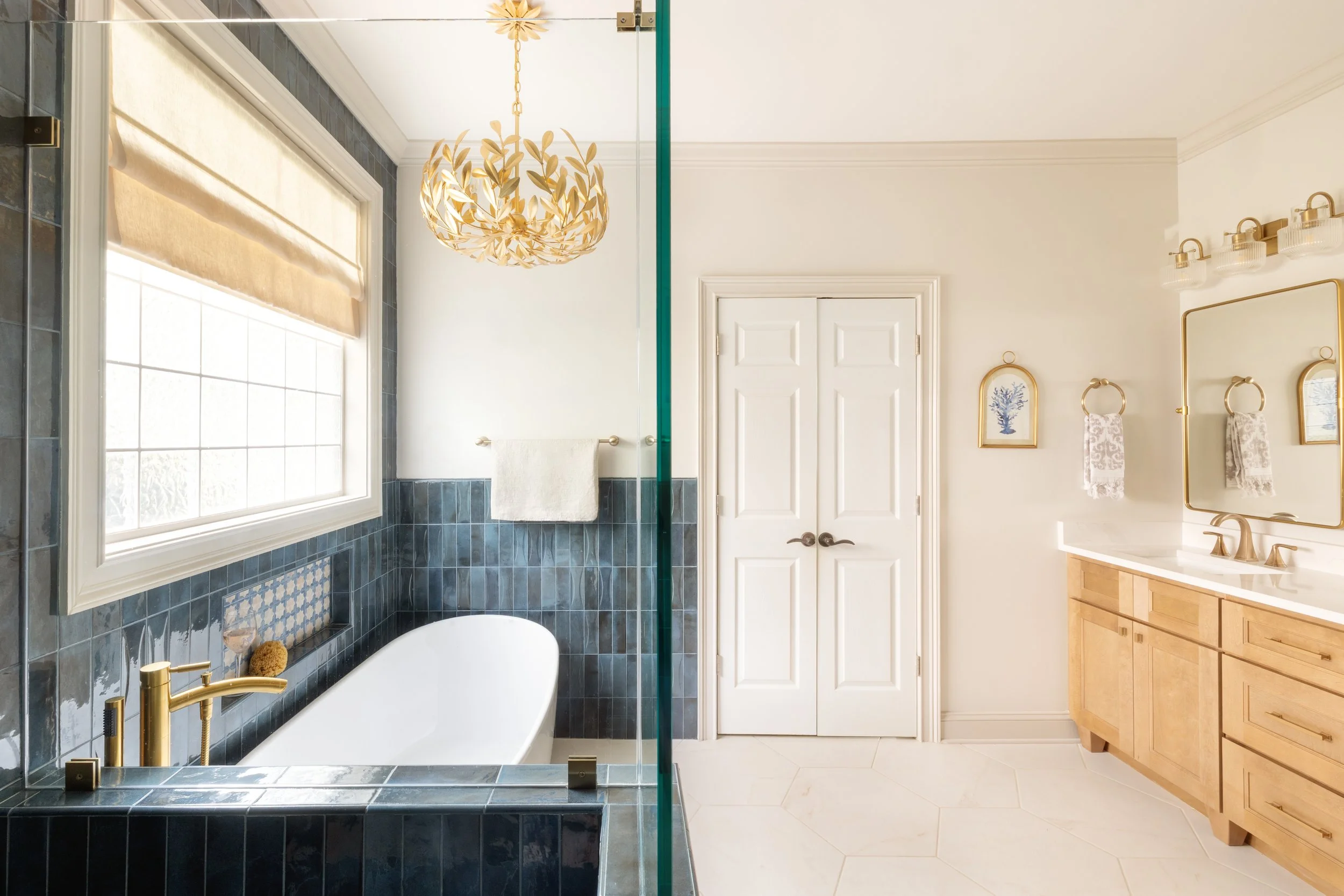 A bathroom with a white bathtub, dark blue tiled walls, a window with a beige Roman shade, a gold faucet, and a gold towel bar with a white towel. There is a decorative paper chandelier hanging from the ceiling. On the right side, there's a white doo
