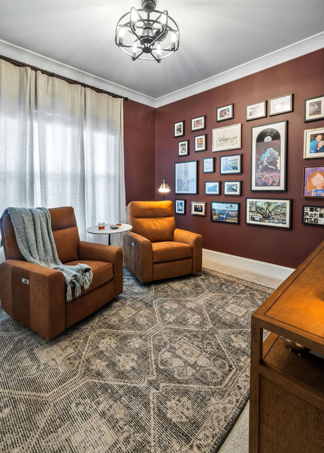 Living room with brown armchairs, a gallery wall of various framed photos and artwork on a maroon wall, a patterned area rug, and a wooden console table.