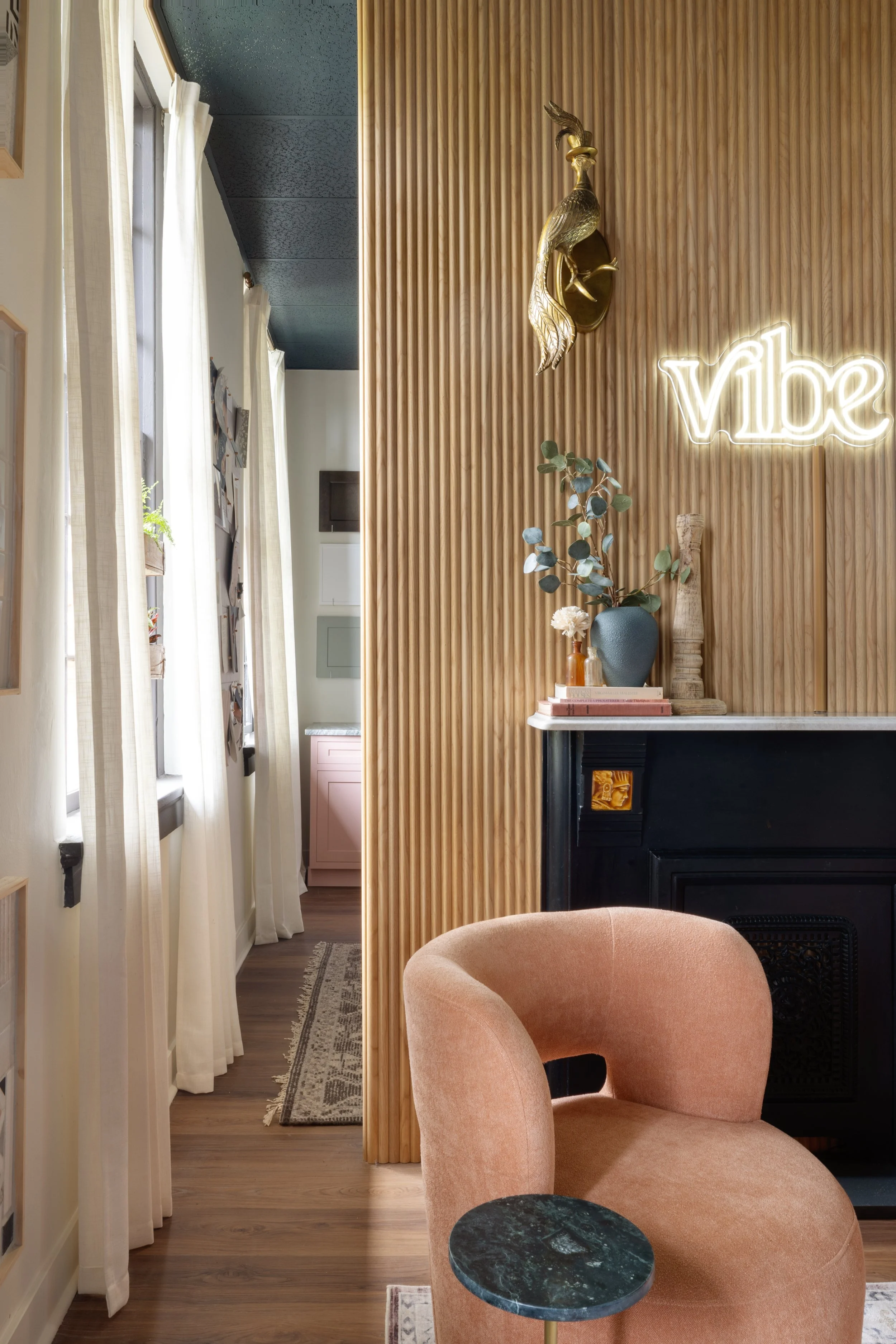 Interior of a cozy living space with a peach upholstered chair, a small green marble side table, a decorative wooden sculpture, a vase with eucalyptus leaves and flowers, and a neon sign that says 'vibe'. The space features a wood-paneled wall with a golden peacock sculpture and natural light coming through sheer curtains.