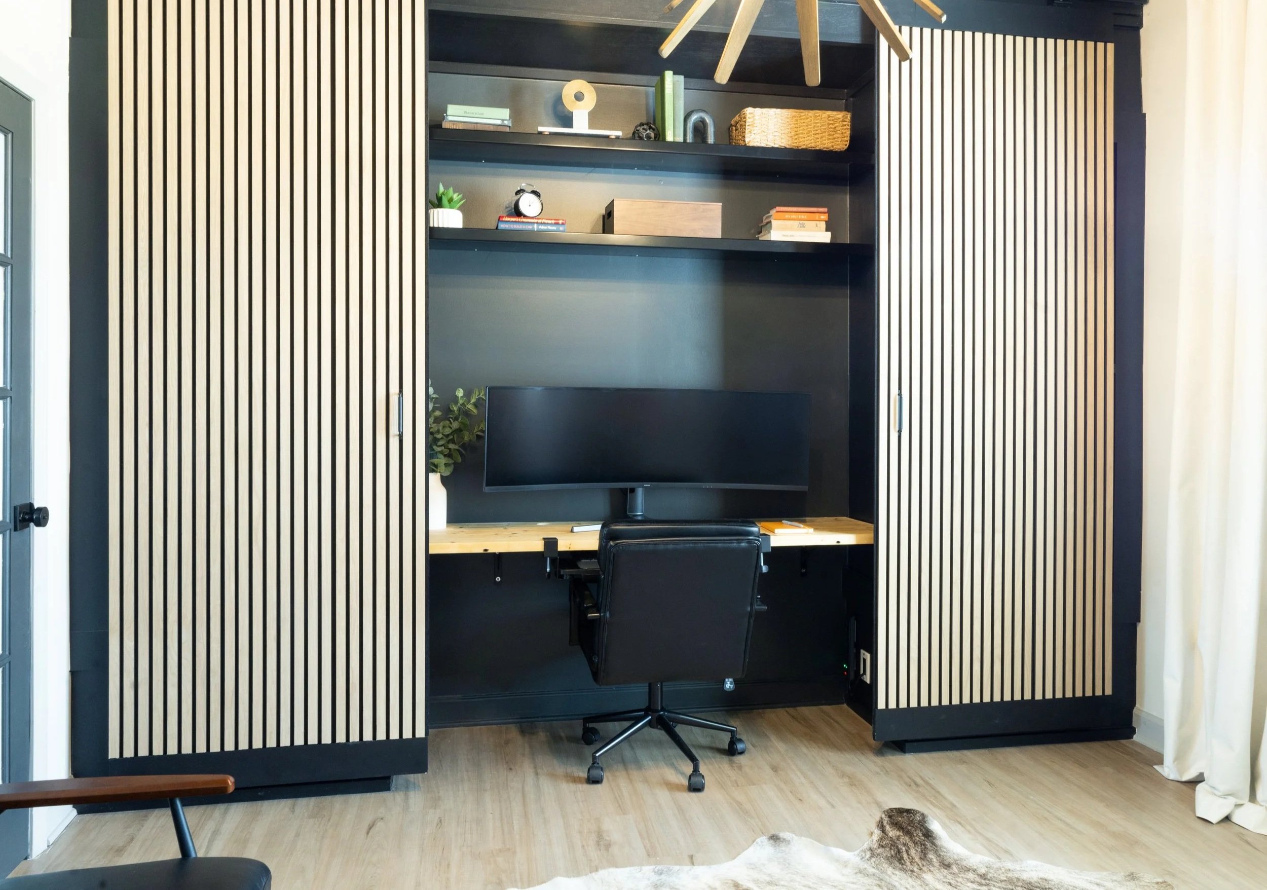 A modern workspace with a black and wood slatted sliding door open to reveal a small office nook. Inside there is a black wall, a wooden desk, a black rolling chair, and three-step shelves holding books, decor items, and a small potted plant. A black monitor is on the desk, with a cowhide rug on the light wood floor in front of it.