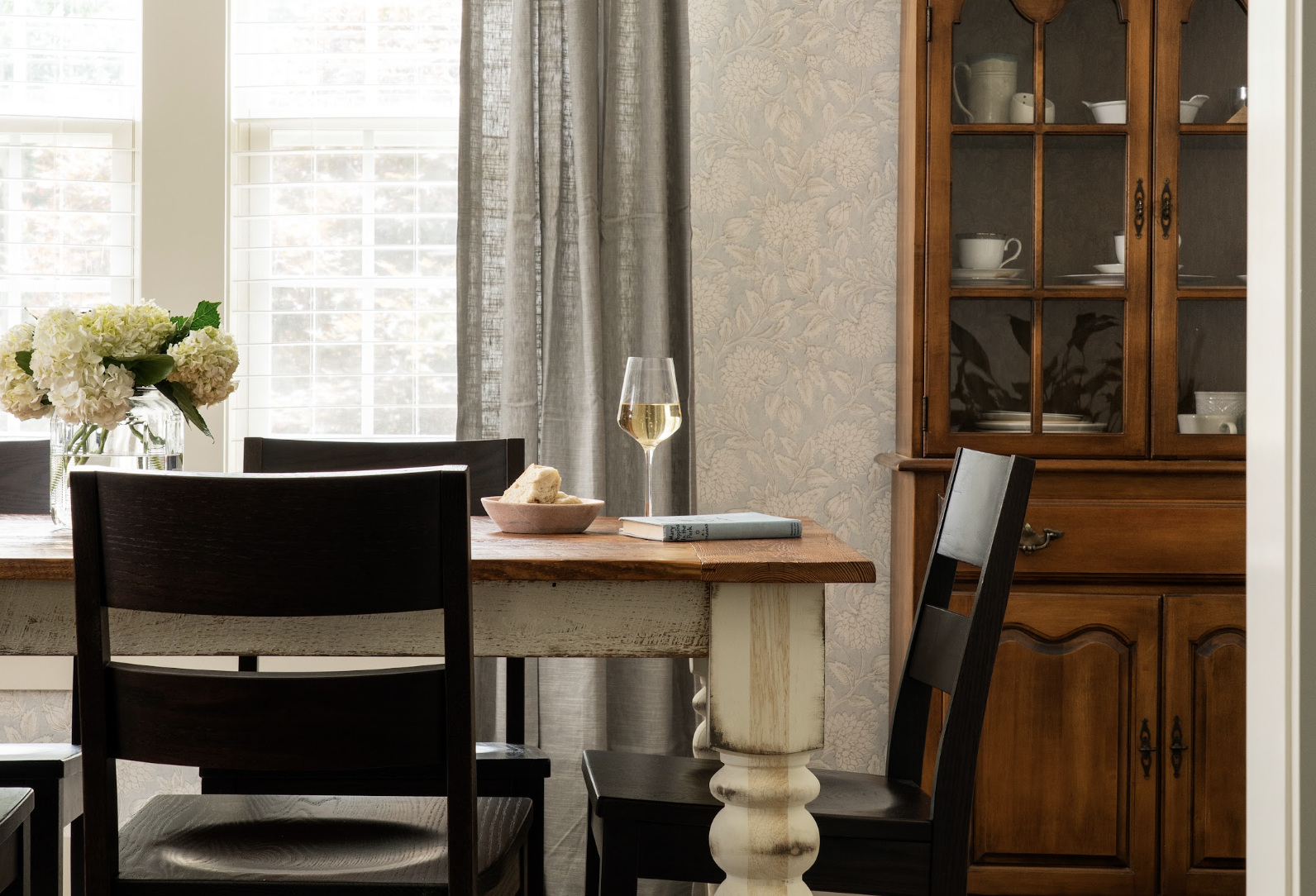 A dining room with a wooden table set with a vase of white flowers, a glass of white wine, a bowl with bread, and a book. There are black chairs around the table and a wooden china cabinet with dishes inside. Large windows with white blinds and gray curtains are in the background.