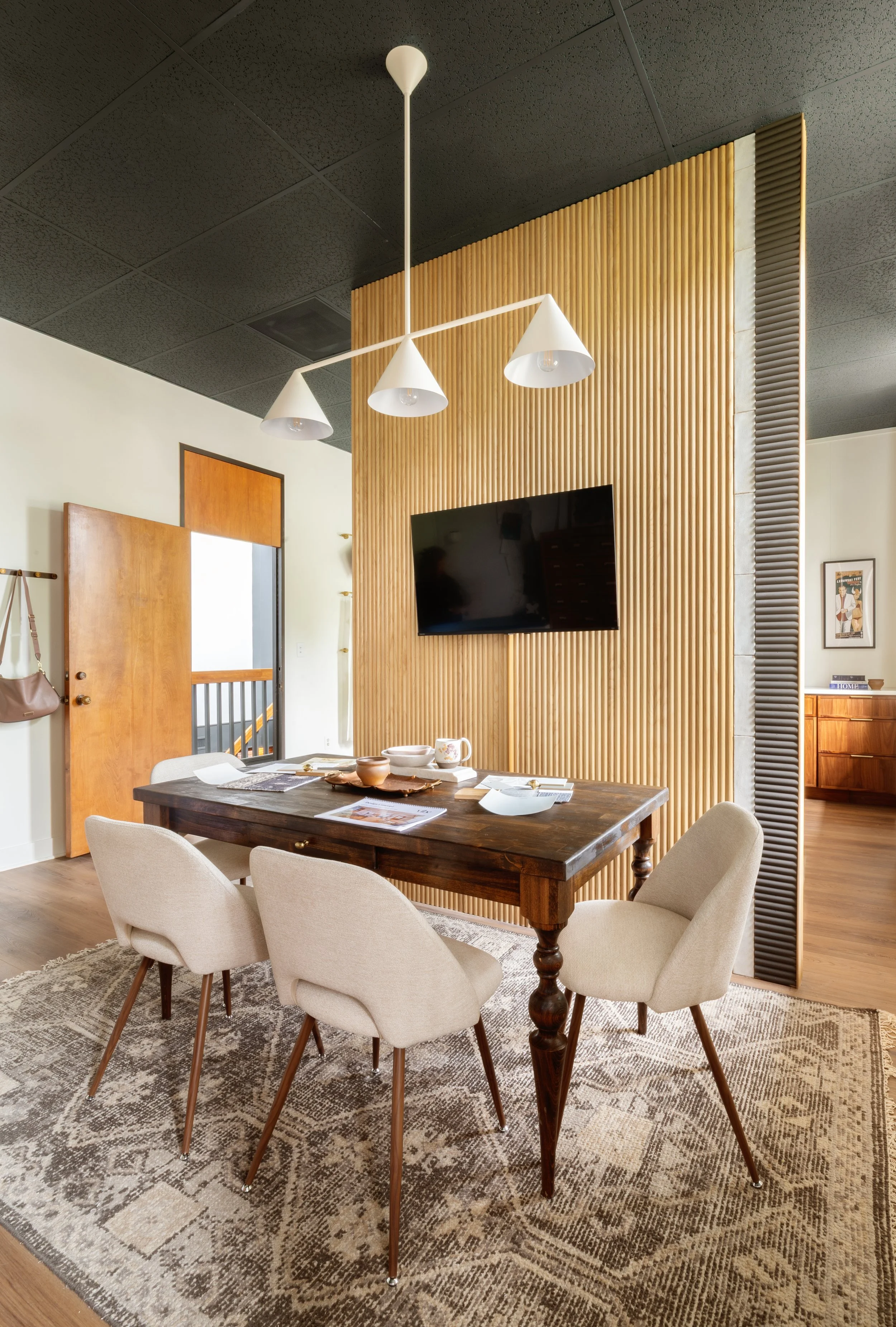 A dining room with a wooden rectangular table surrounded by six beige upholstered chairs, a patterned rug underneath, a hanging white pendant light fixture, a wood paneled wall with a flat-screen TV, and a doorway leading to a staircase area.
