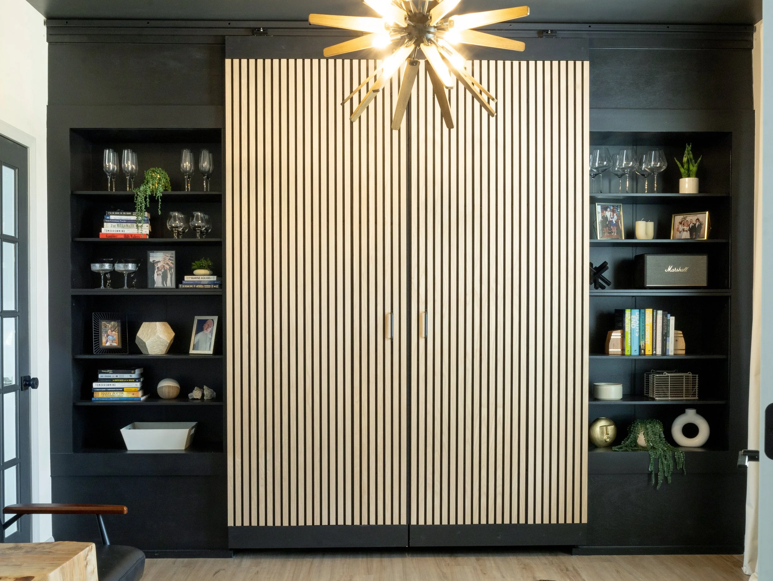 A black cabinet with striped wooden sliding doors in a room. The cabinet shelves hold glassware, picture frames, books, a small plant, and decorative objects. A modern chandelier with wooden blades hangs from the ceiling.