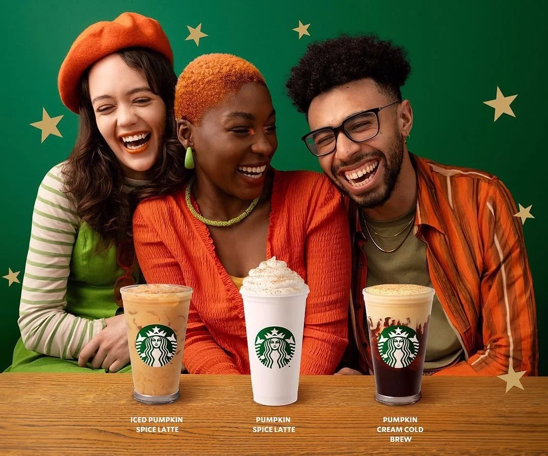 Three people smiling and laughing at a table with three Starbucks drinks: iced pumpkin spice latte, pumpkin spice latte, and pumpkin cream cold brew, set against a green background with star decorations.
