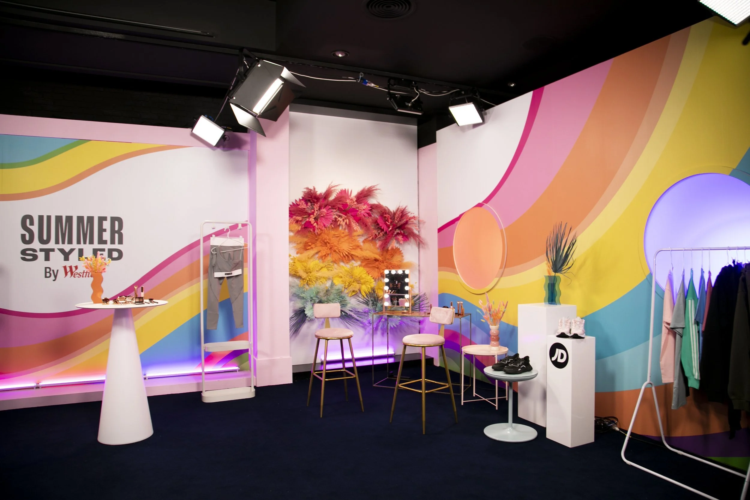 Colorful room with pink, yellow, orange, green, blue, and purple abstract wall art, mannequins with clothing and shoes, chairs, vases with decorative plants, a mirror with lights, and a logo that says "Summer Styled by Westfield."