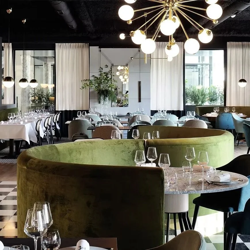 L'intérieur d'un restaurant. Au premier plan un canapé en velour vert, des tables dressées avec des couvert et des verres. Lieu élégant avec une décoration noire, doré, et des matières noble comme le bois et du marbre.