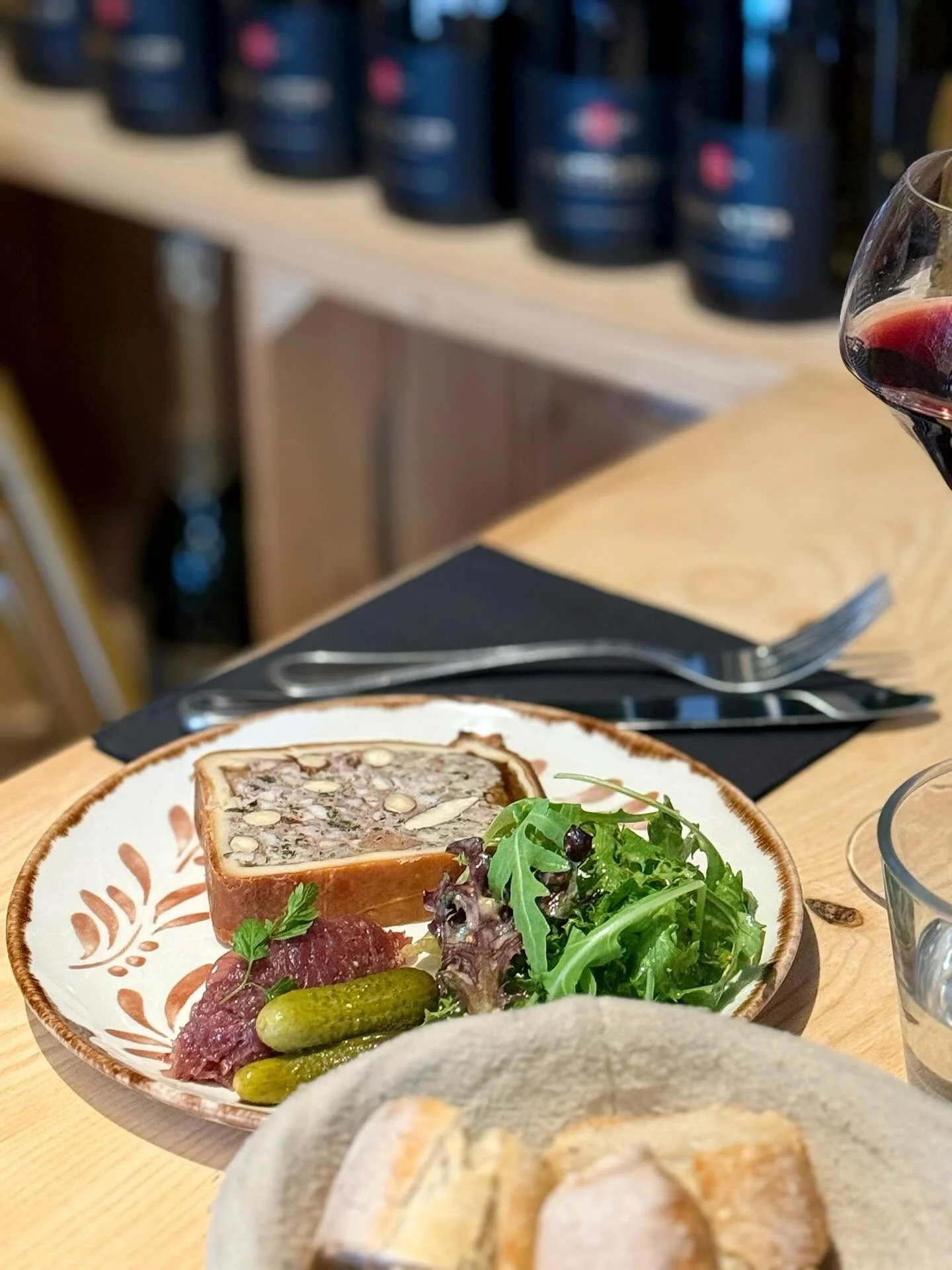 Assiette en grès avec une tranche de pâté en croute, des cornichons, une salade, servi avec un verre de vin rouge. Accompagné des couverts sur un serviettes, le tout posé sur une table en bois.