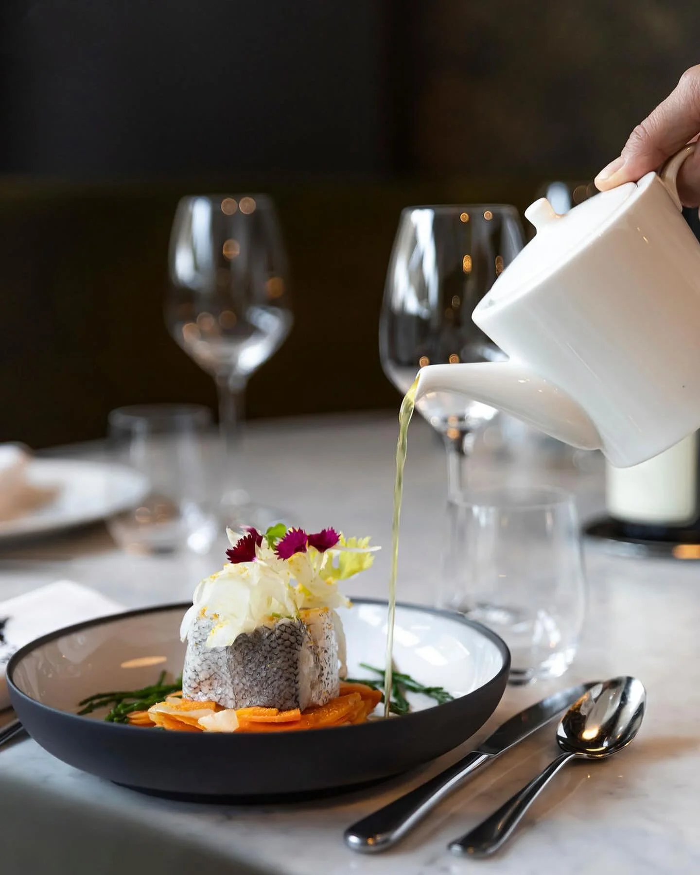 En premier plan une assiette creuse dans laquelle il y a un plat avec des carottes et du poisson. Une main tenant une théière et versant du thé dans l'assiette est au dessus du plat. En arrière plan des verres à pieds.
